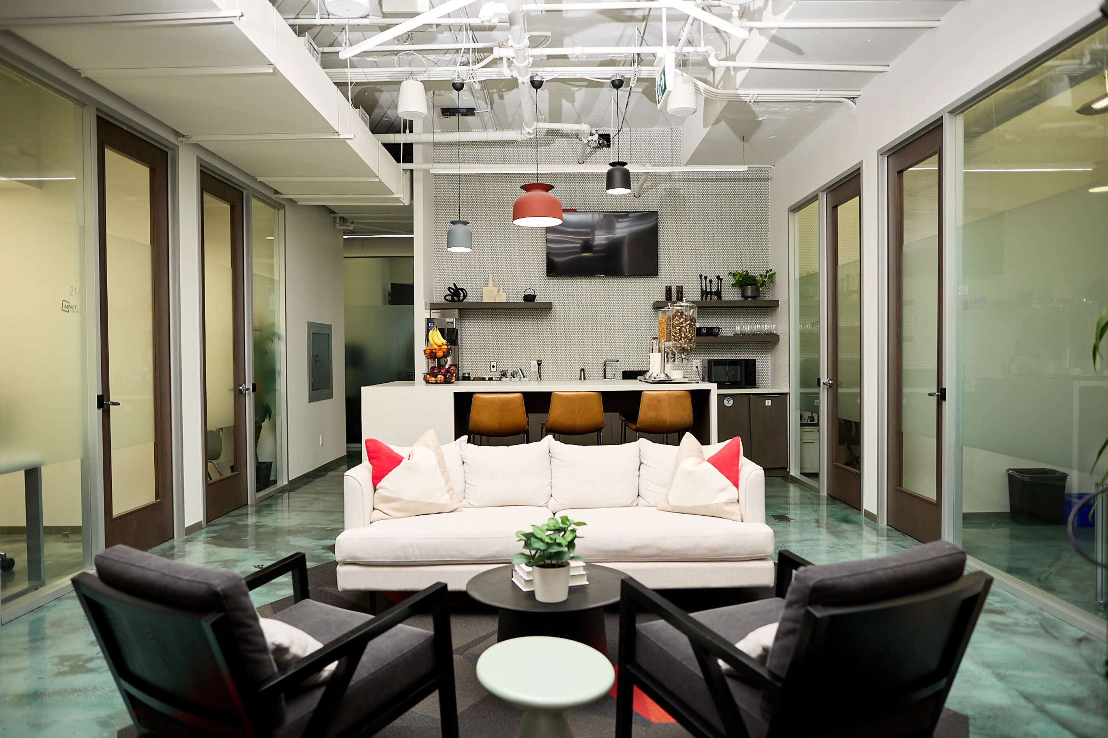 The image shows a modern office lounge area with a white sofa, two black armchairs, and a kitchenette in the background.