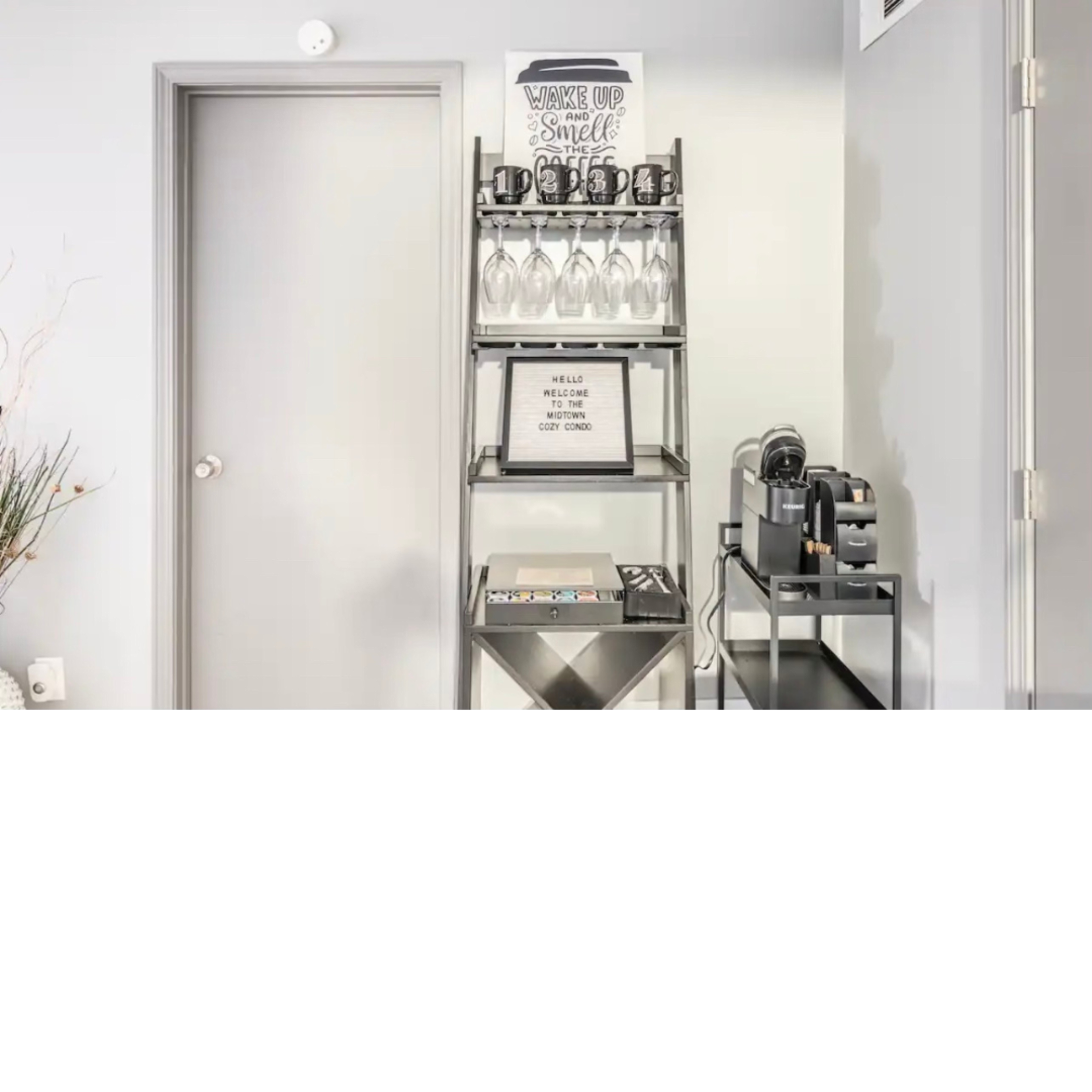 A black metal shelving unit holds glasses and a coffee maker in a well-lit corner of a room.