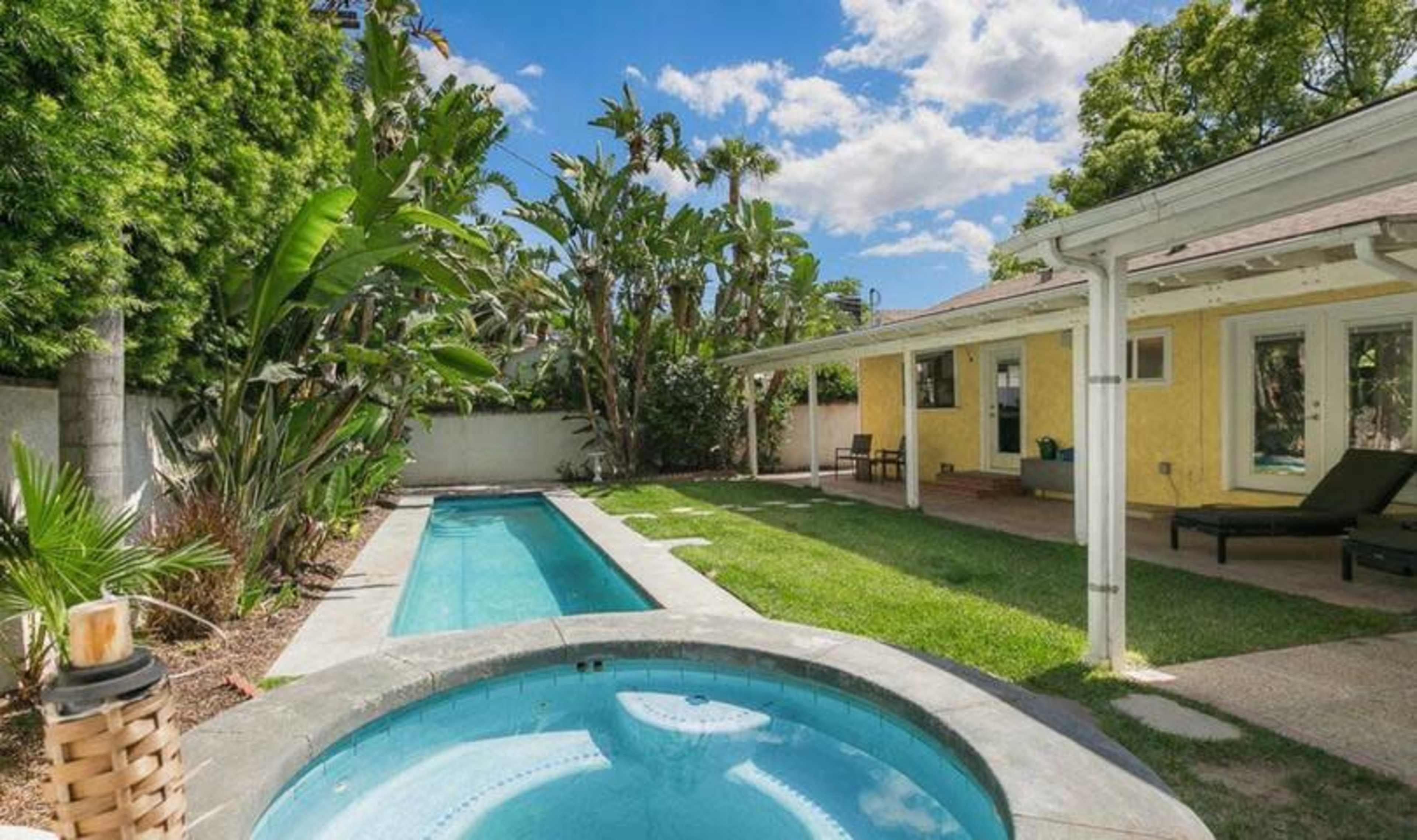 A backyard with a swimming pool, a spa, and a grassy area surrounded by tropical plants and a house with yellow walls.
