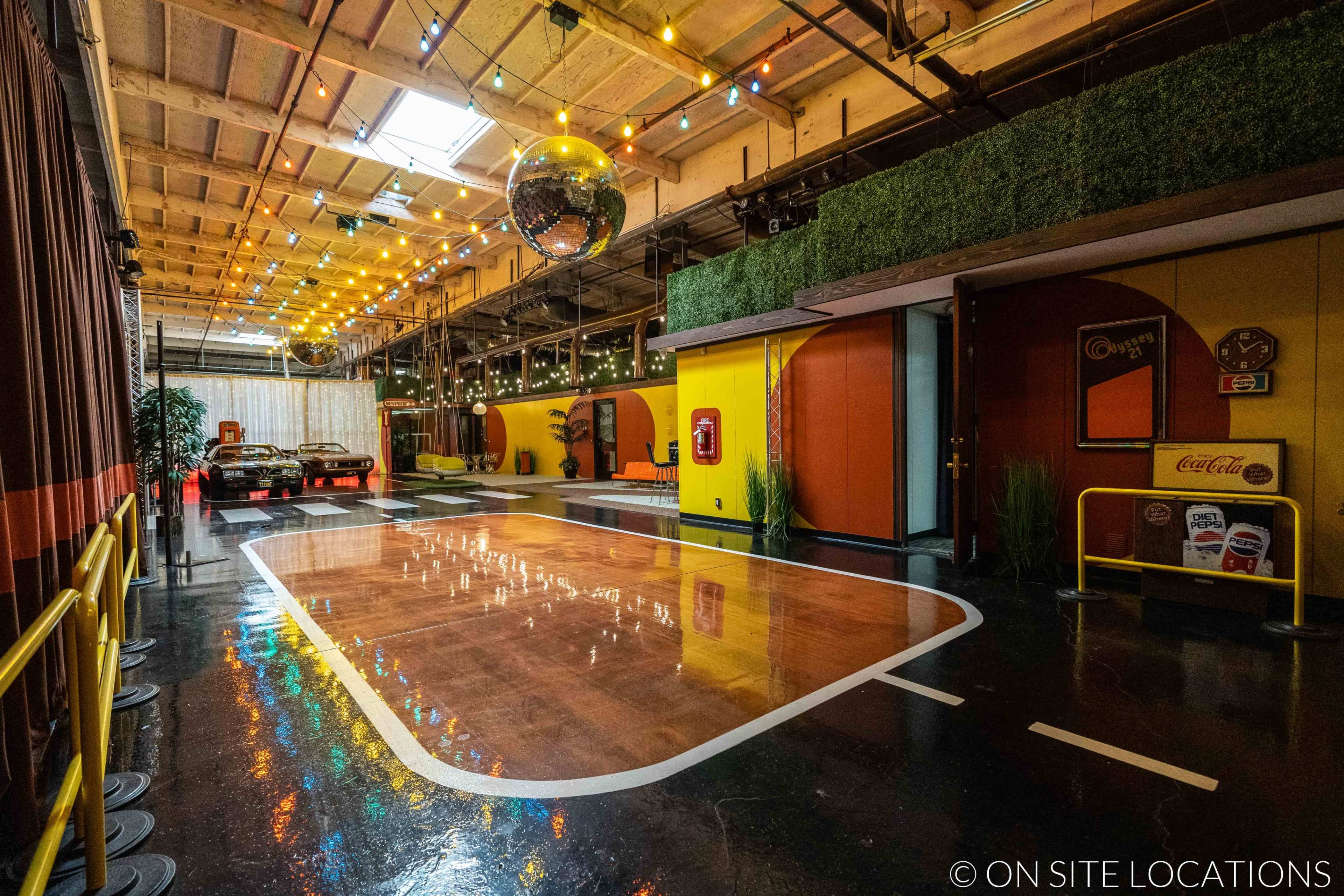 The image shows a vibrant indoor space featuring a polished floor with a racetrack design, colorful walls, and a disco ball overhead, set up in a retro theme with vintage cars and decor elements.