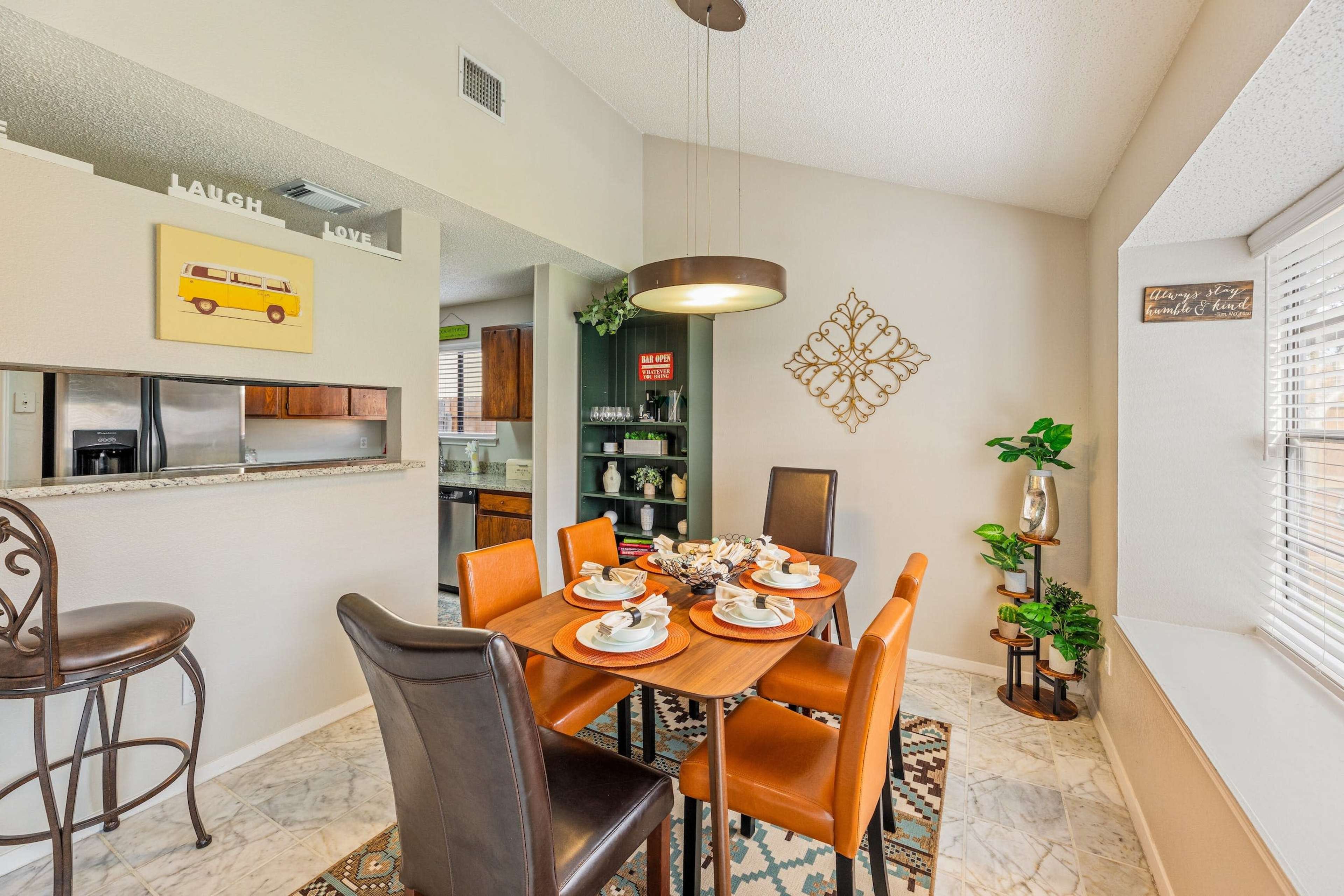 A dining area features a round table with orange and black chairs, set with dishes, surrounded by light-colored walls and a window.