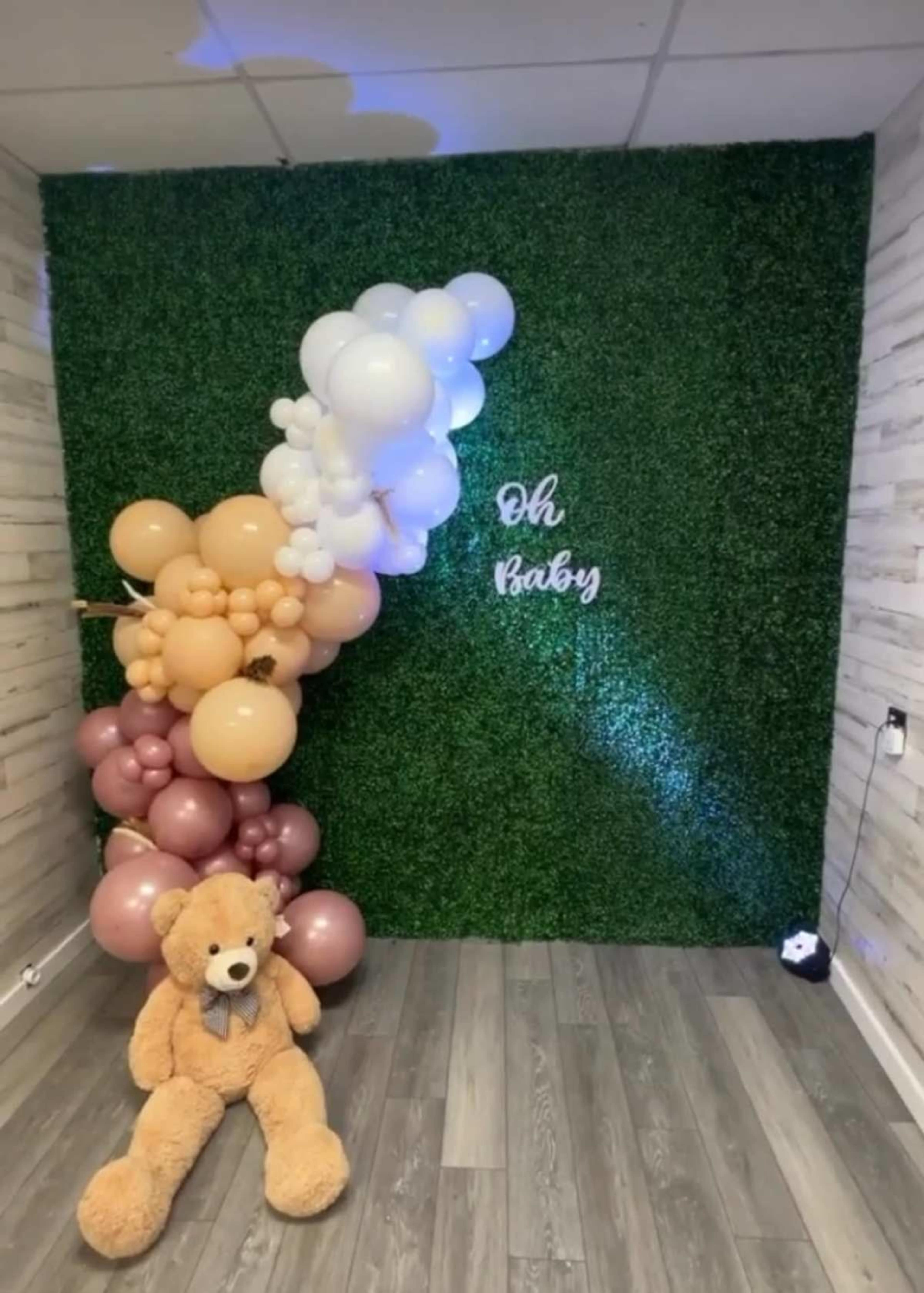 The image shows a decorated wall with green foliage and a balloon arrangement, featuring the words "Oh baby," alongside a plush teddy bear sitting on the floor.