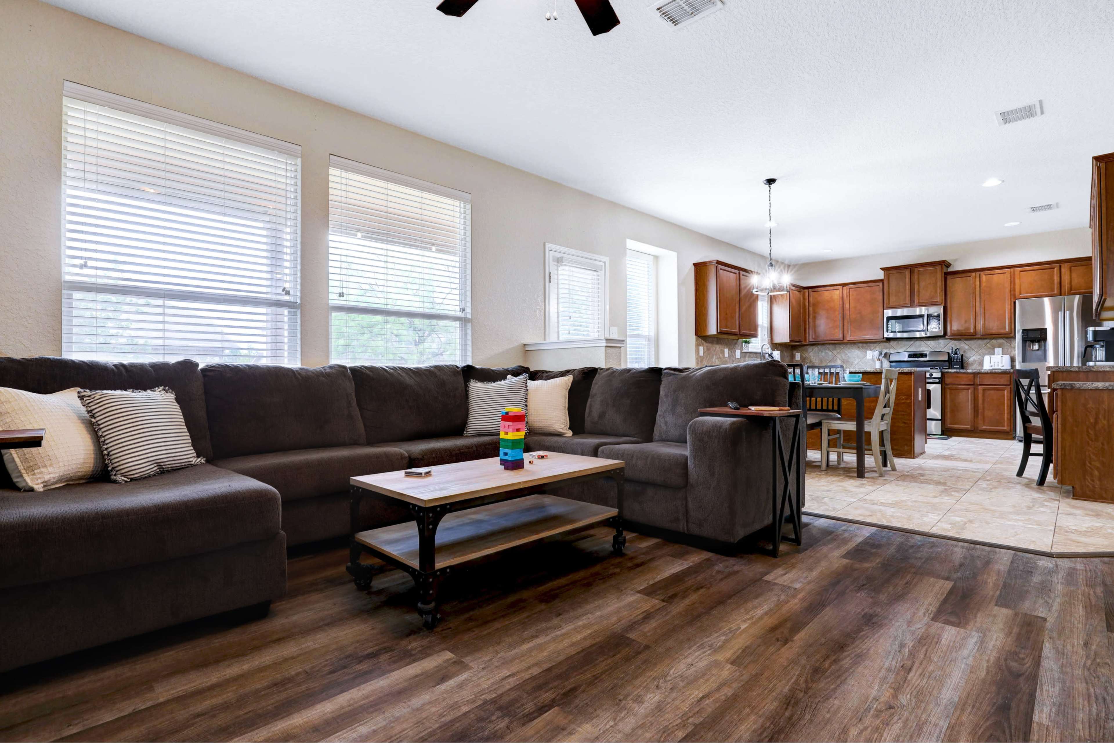 A living room featuring a large gray sectional sofa, a coffee table, and a view into a kitchen with wooden cabinets and stainless steel appliances.