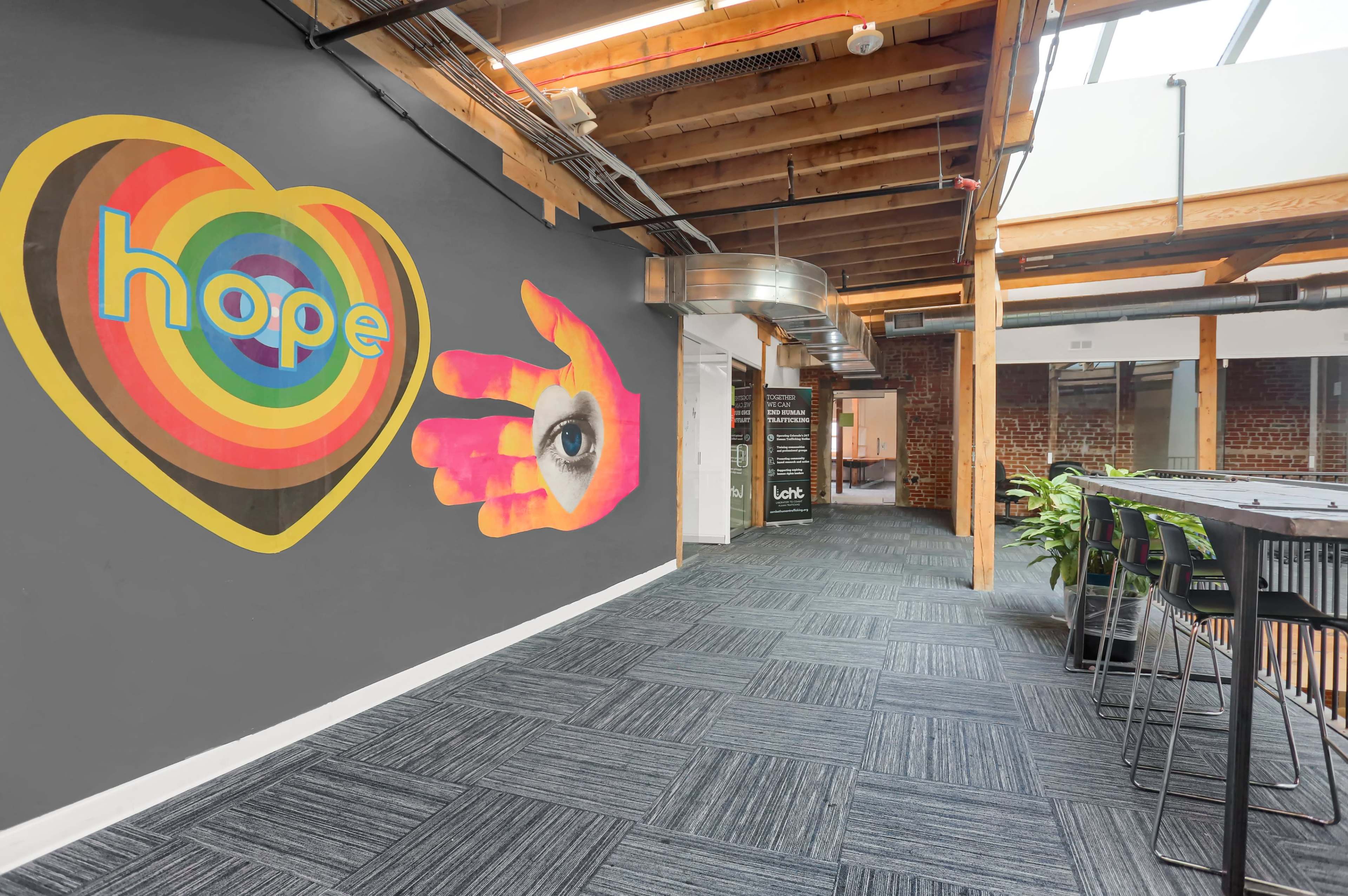 The image shows a modern office space featuring a colorful mural of a heart with the word "hope" and a hand with an eye, alongside an open hallway with a seating area and exposed wooden beams.