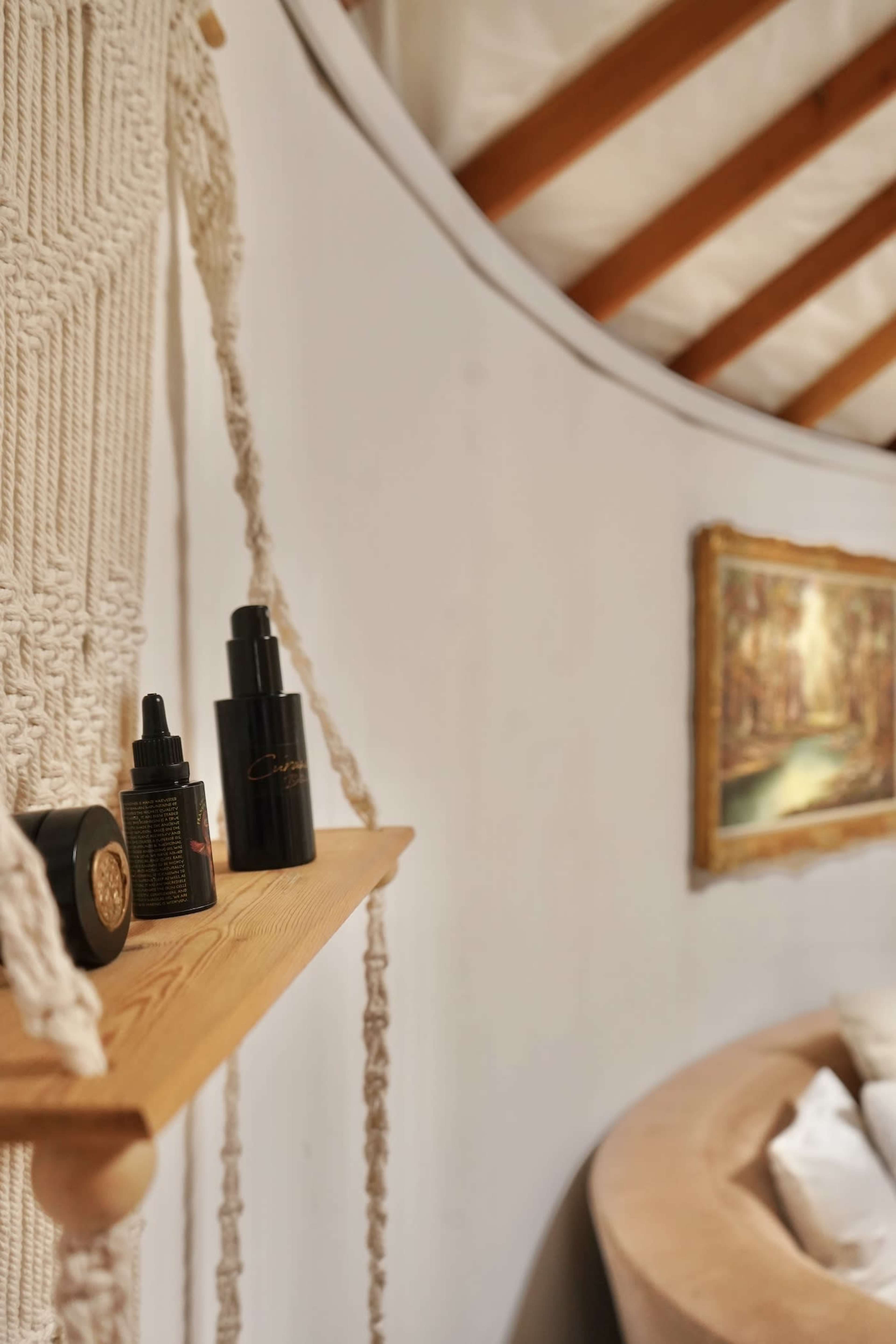 A wooden shelf with various skincare bottles against a curved white wall, with a painting of a landscape hanging nearby.