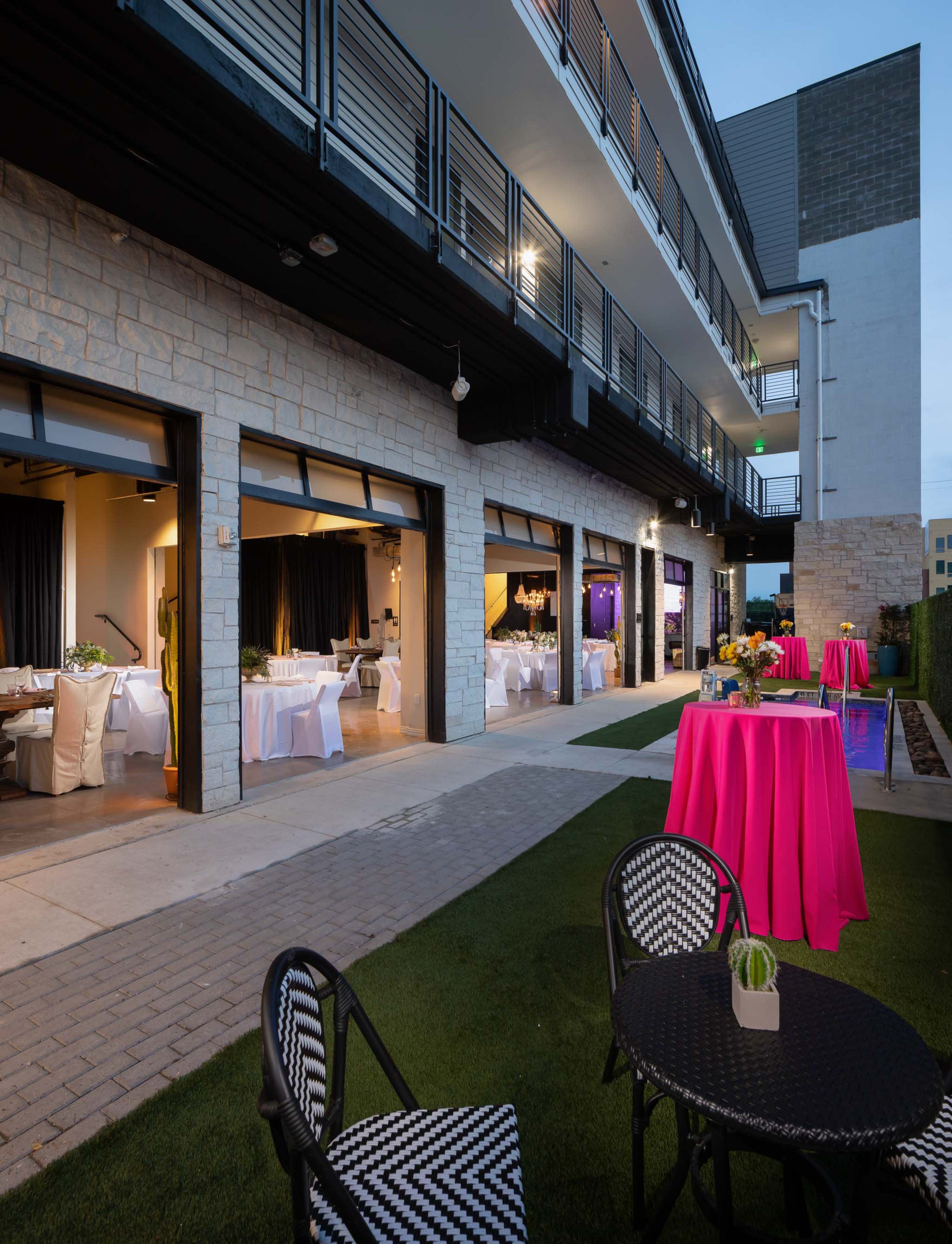 An outdoor area with pink tablecloths and chairs, adjacent to a building with large open doors leading to elegantly set dining spaces.