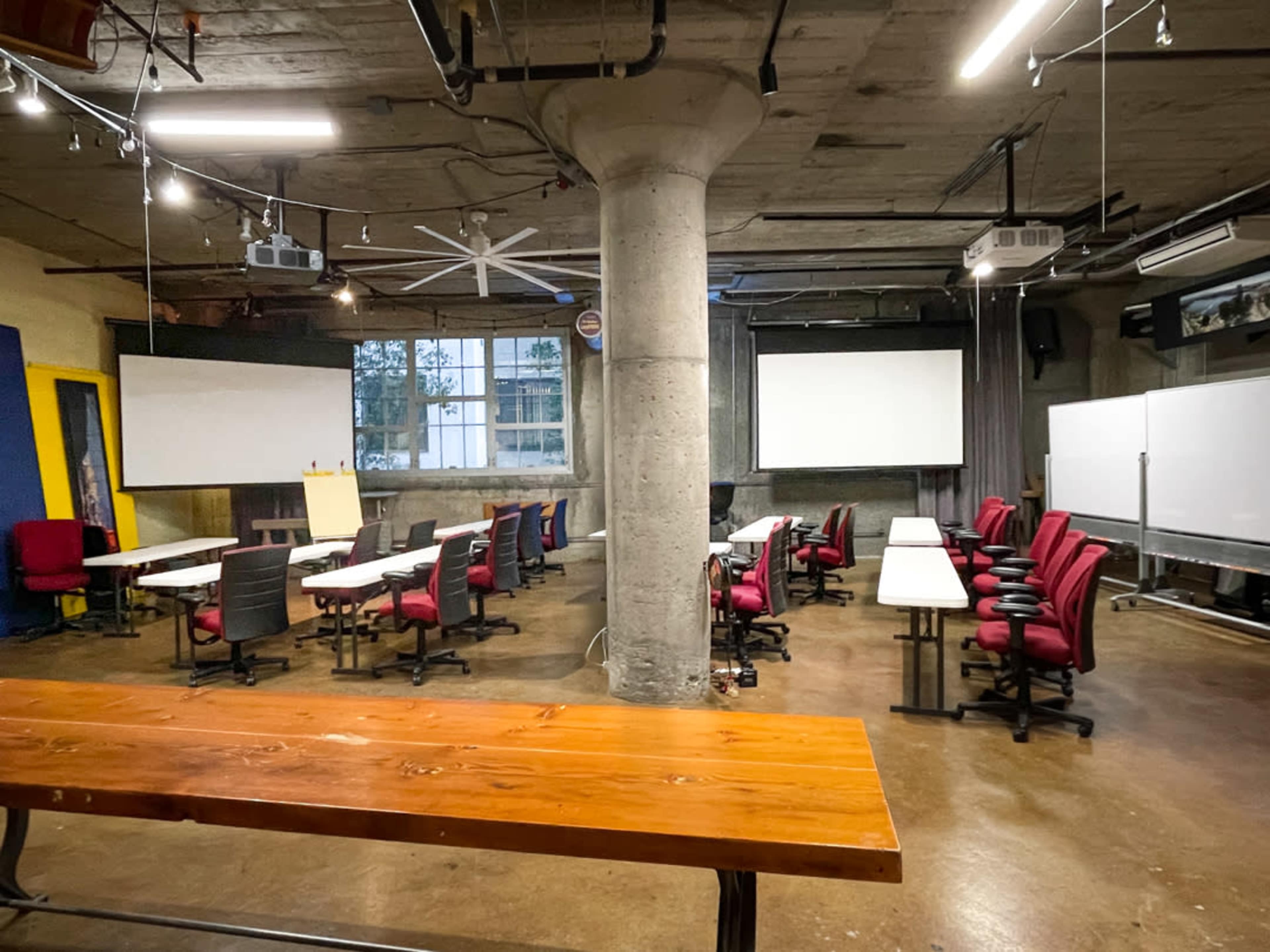 A spacious classroom features rows of red chairs and white tables, with large projection screens mounted on the walls.