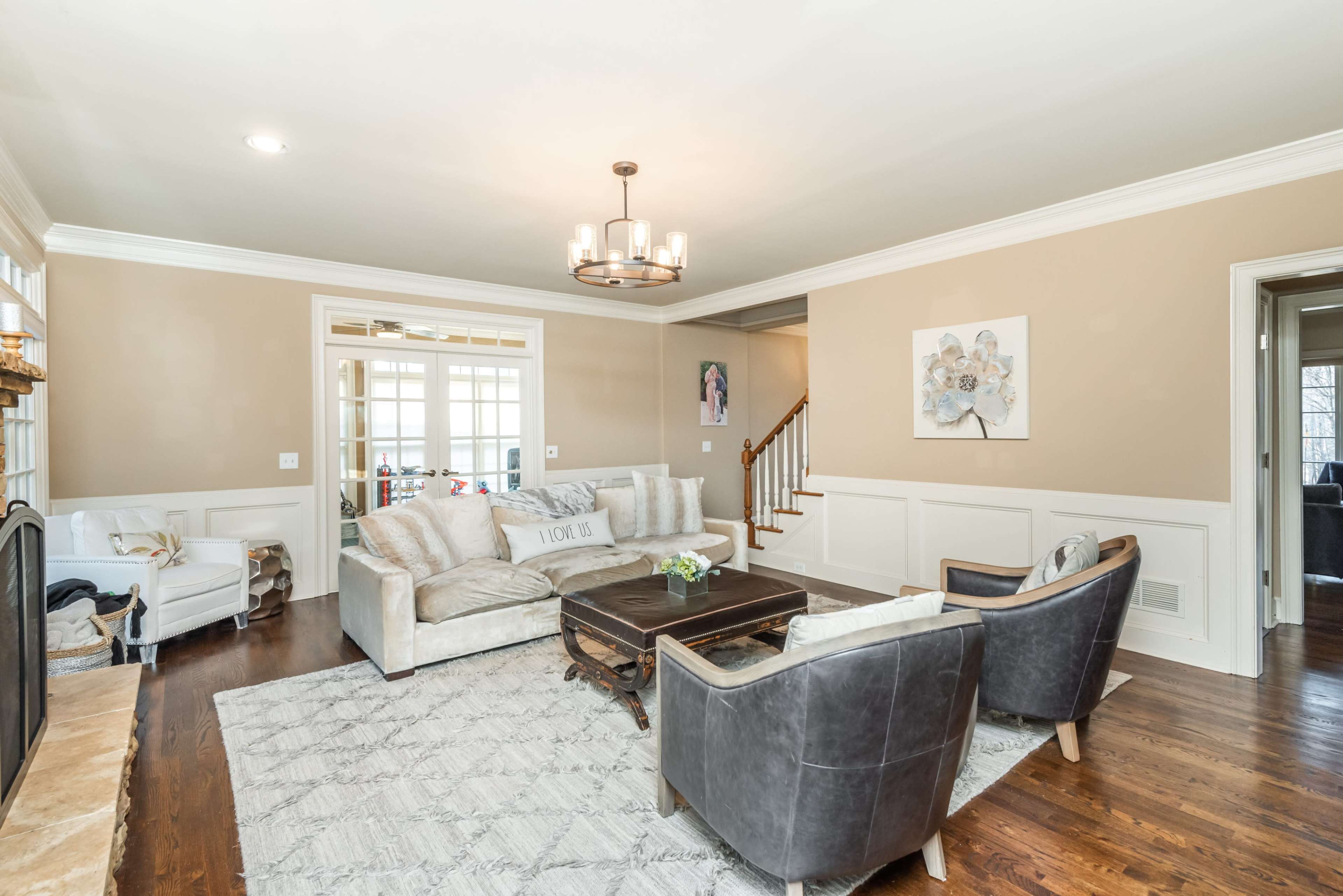 A light-filled living room features a beige sofa, dark chairs, a wooden coffee table, and a decorative wall with wainscoting.
