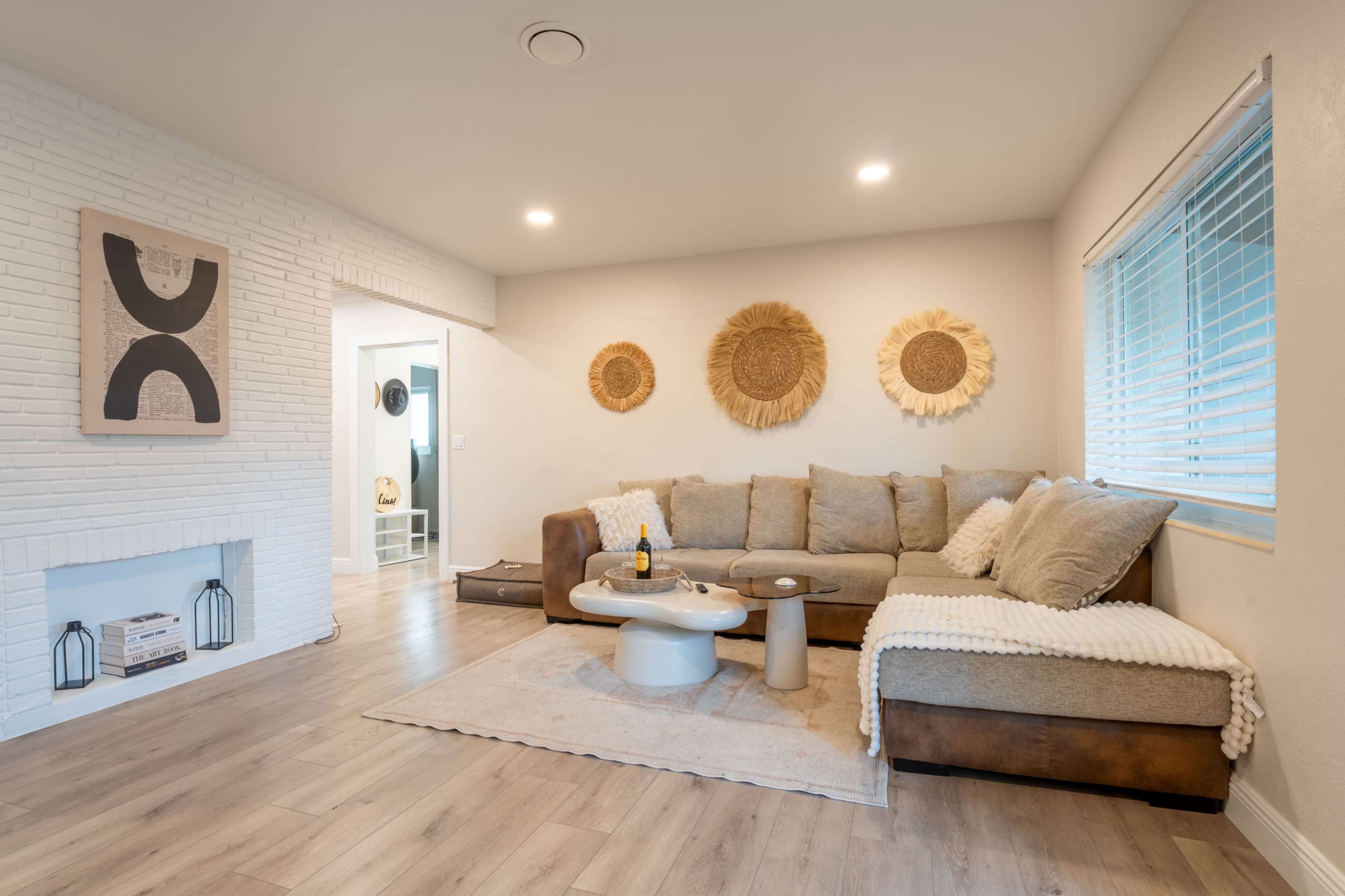 The image shows a cozy living room with a large sectional sofa, a round coffee table, and decorative wall hangings, featuring a light color scheme and wooden flooring.