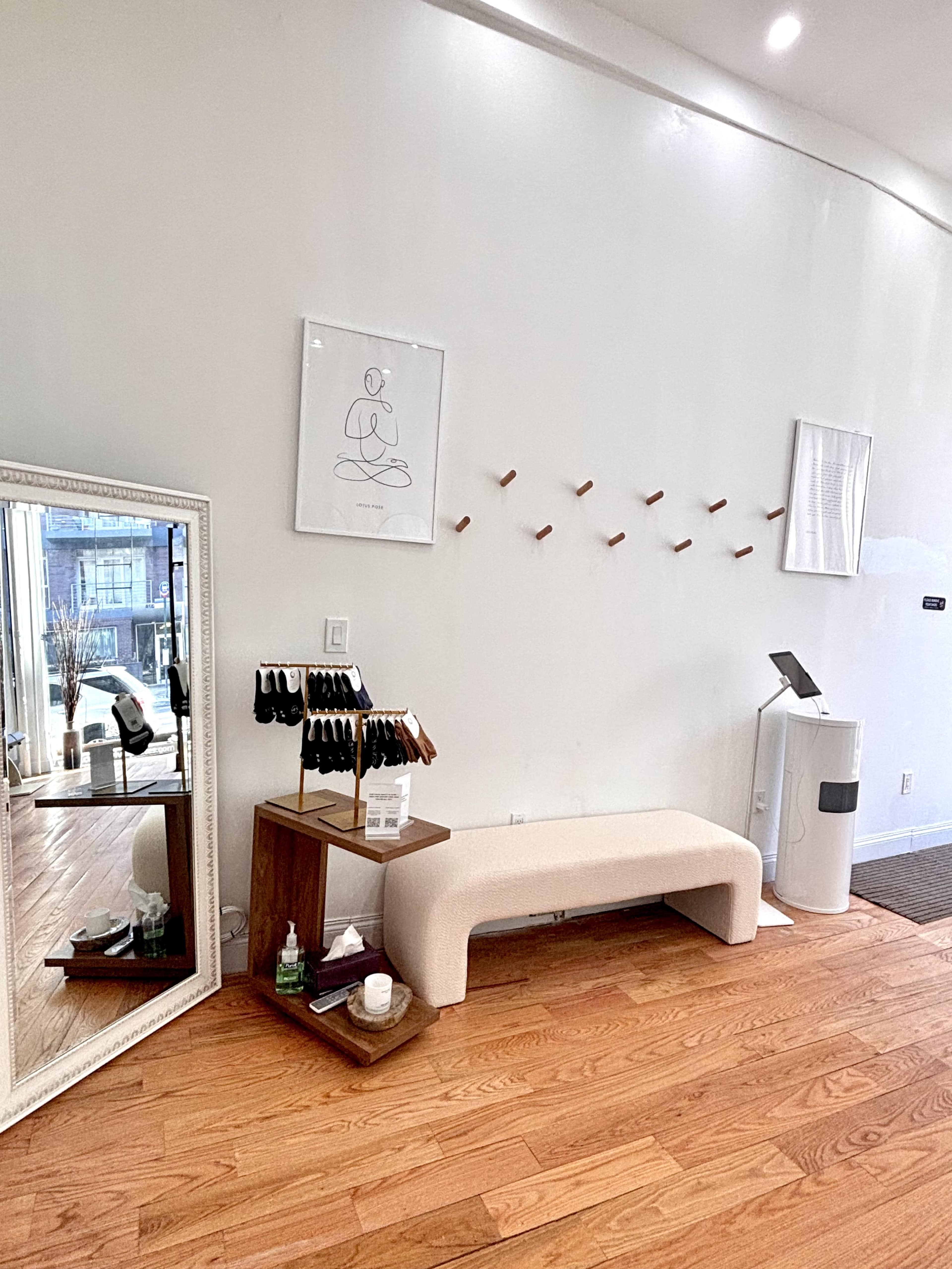 The image shows a minimalist interior of a wellness space, featuring a bench, mirror, and decorative wall hangings.