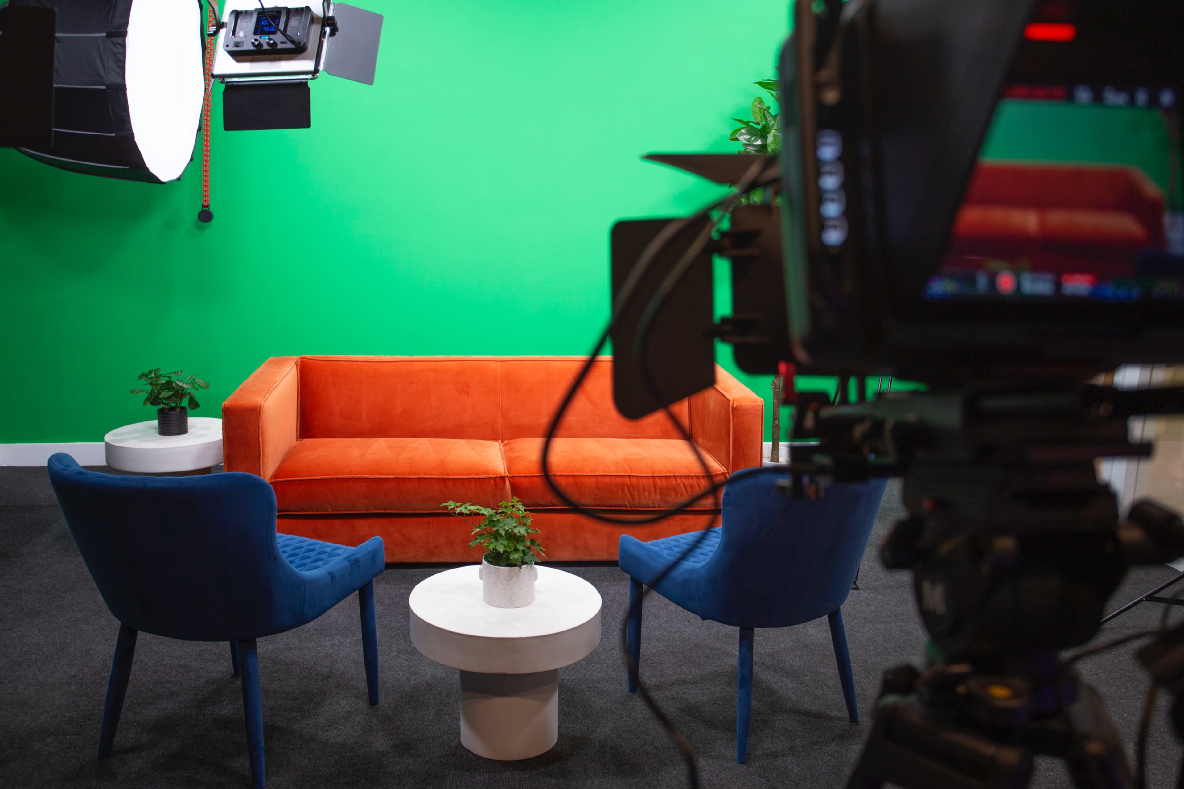 A television studio setup with an orange couch, two blue chairs, and a small white table, all in front of a green screen.