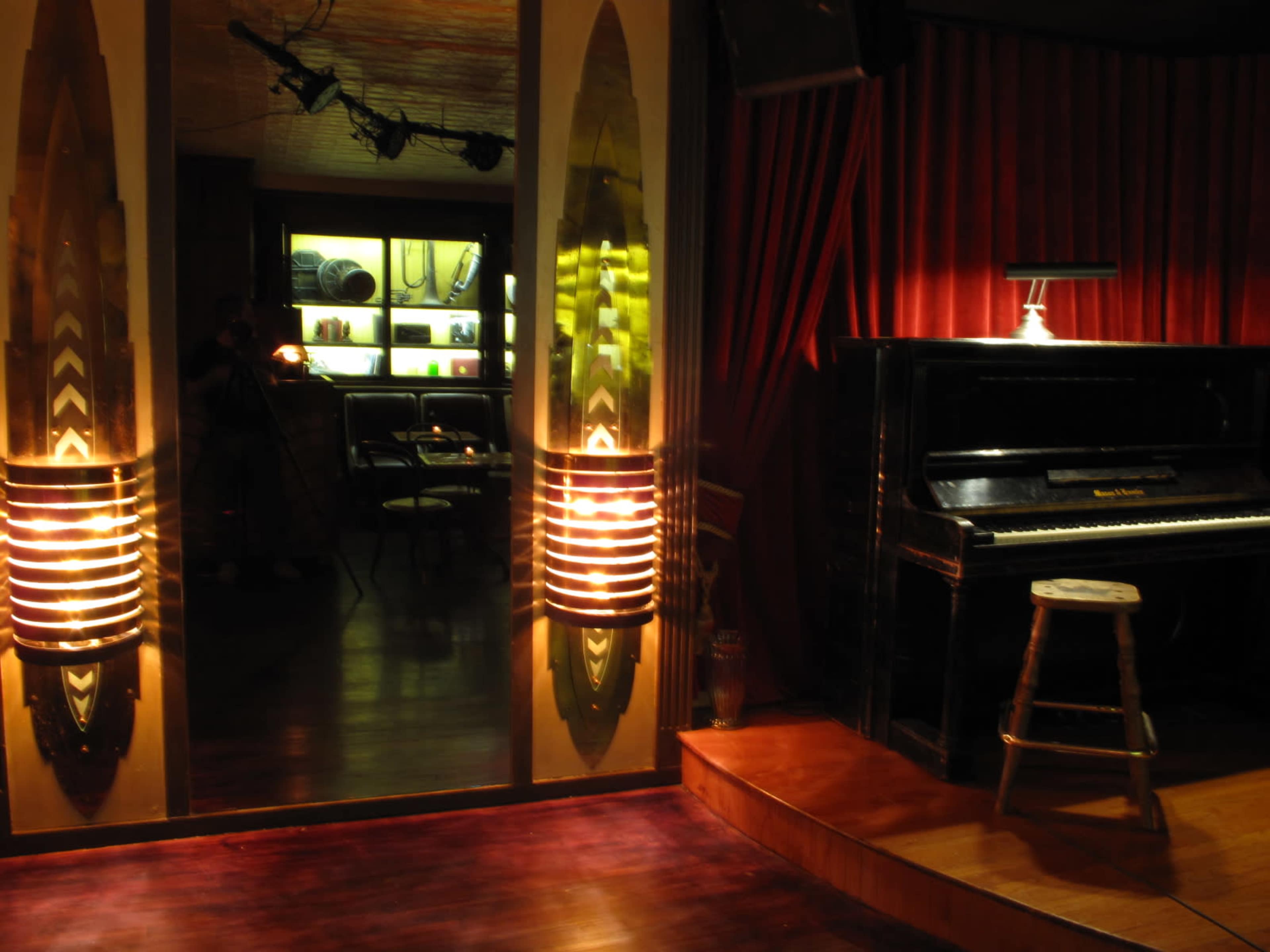 The image depicts a dimly lit room featuring a grand piano beside a stool, with a red curtain in the background and a reflective surface showcasing items on shelves.