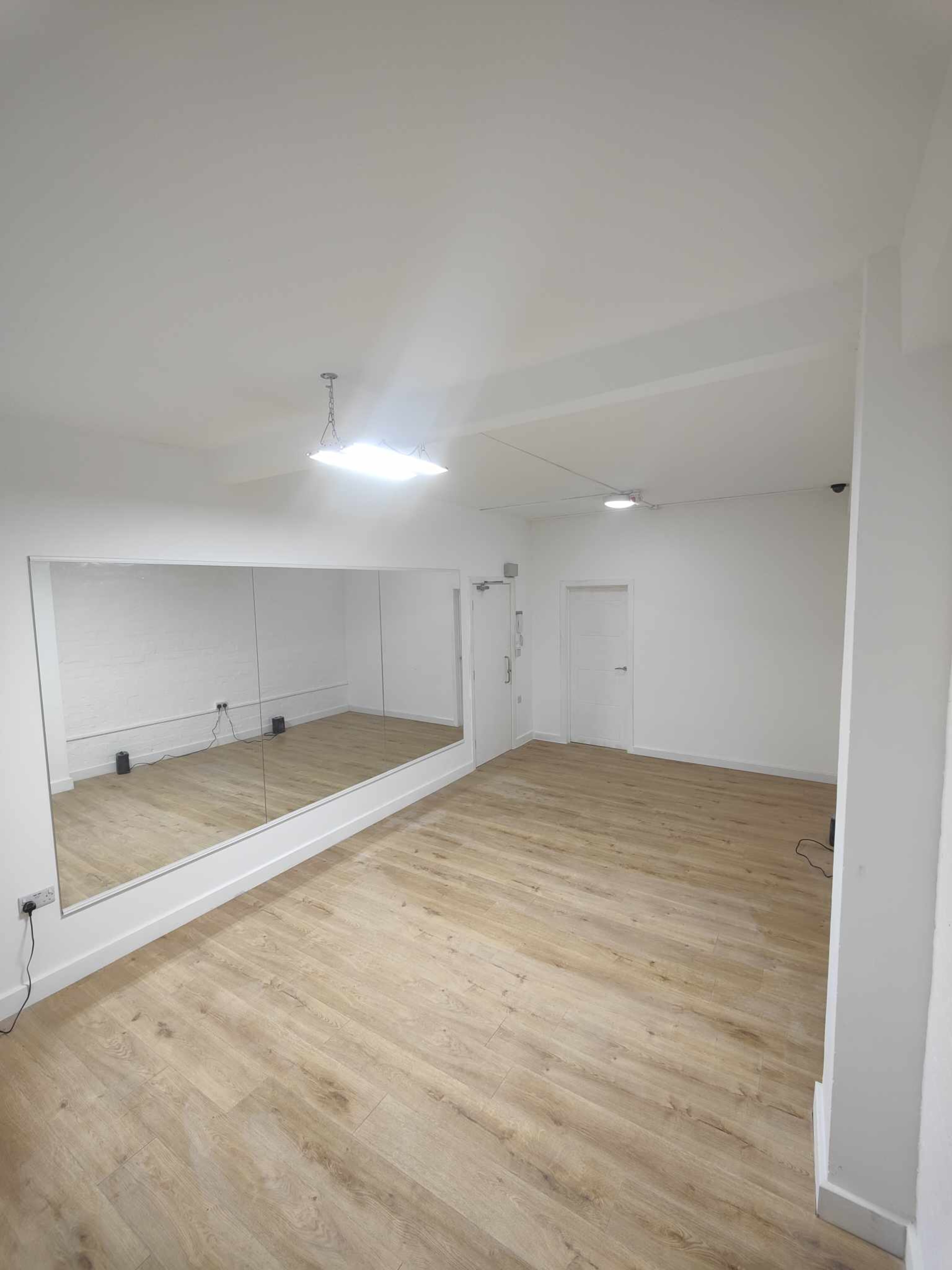 The image shows a spacious, well-lit room with wooden flooring and a large mirror on one wall.