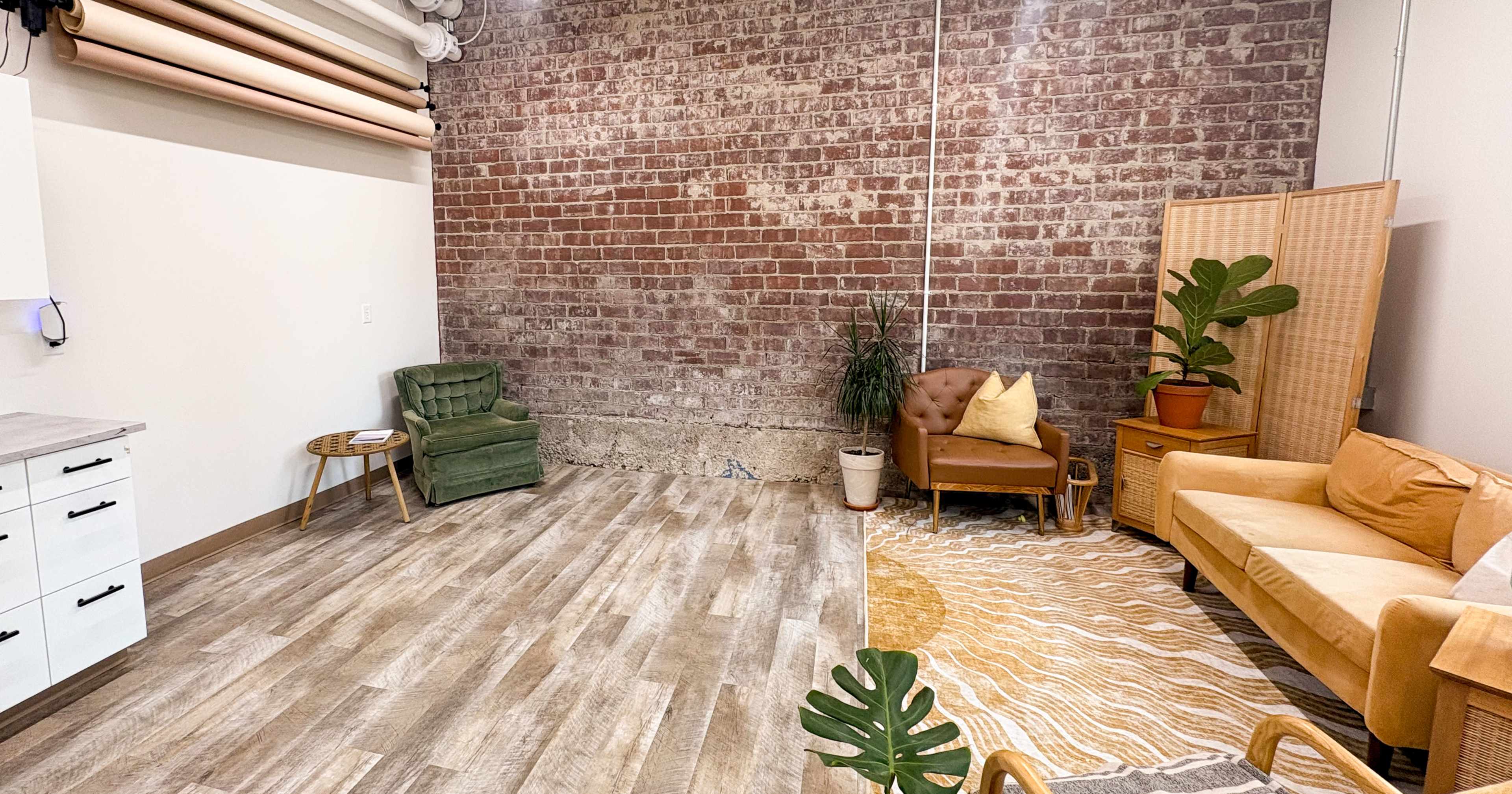 A cozy room features a brick wall, two seating areas with green and orange sofas, a plant, and wooden flooring.