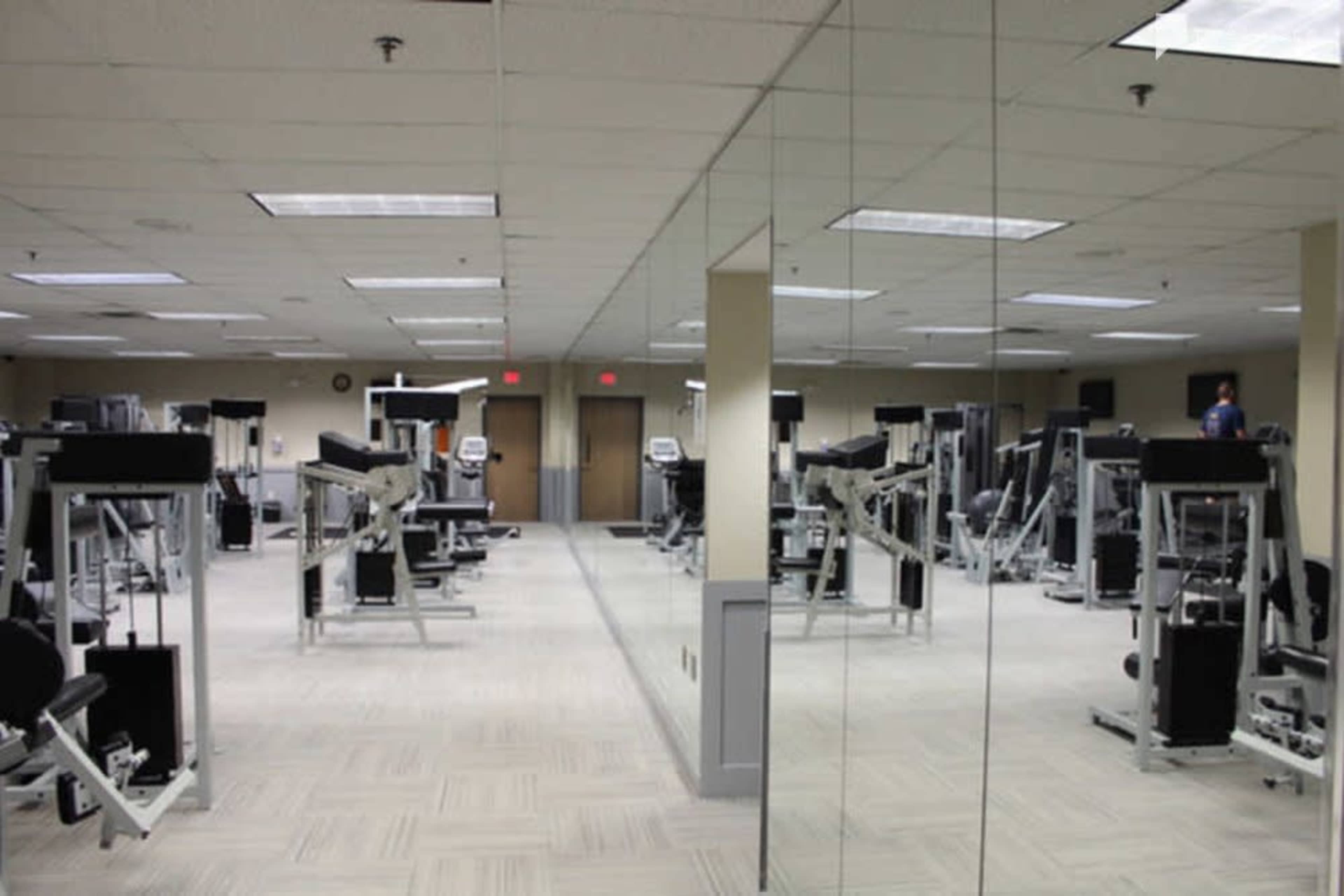 The image shows a gym interior with various exercise machines arranged in two symmetrical rows, reflected in large mirrors on one wall.
