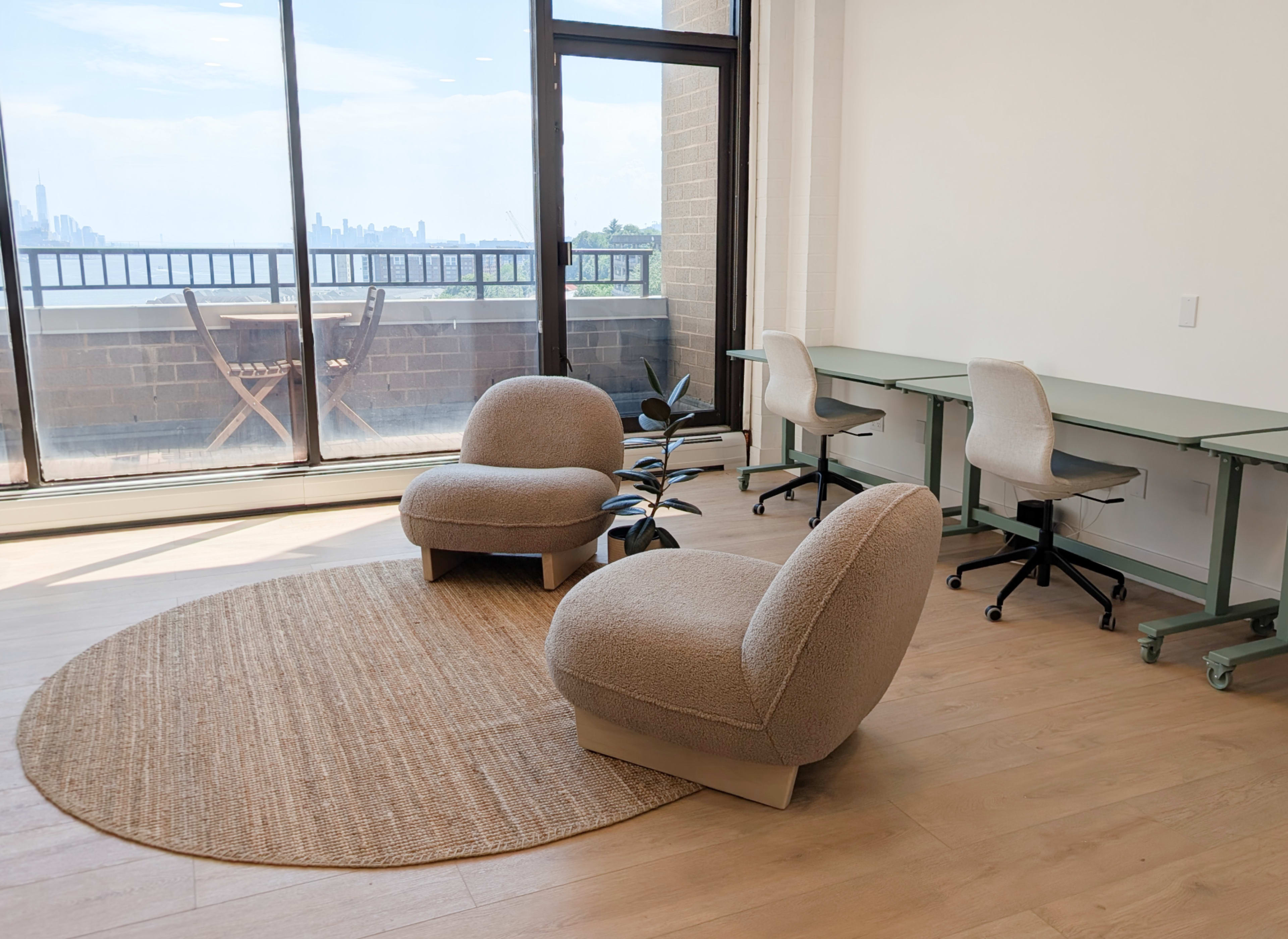 A bright, modern workspace features two cozy chairs on a round rug, with a view of the city skyline through large windows and a desk area nearby.