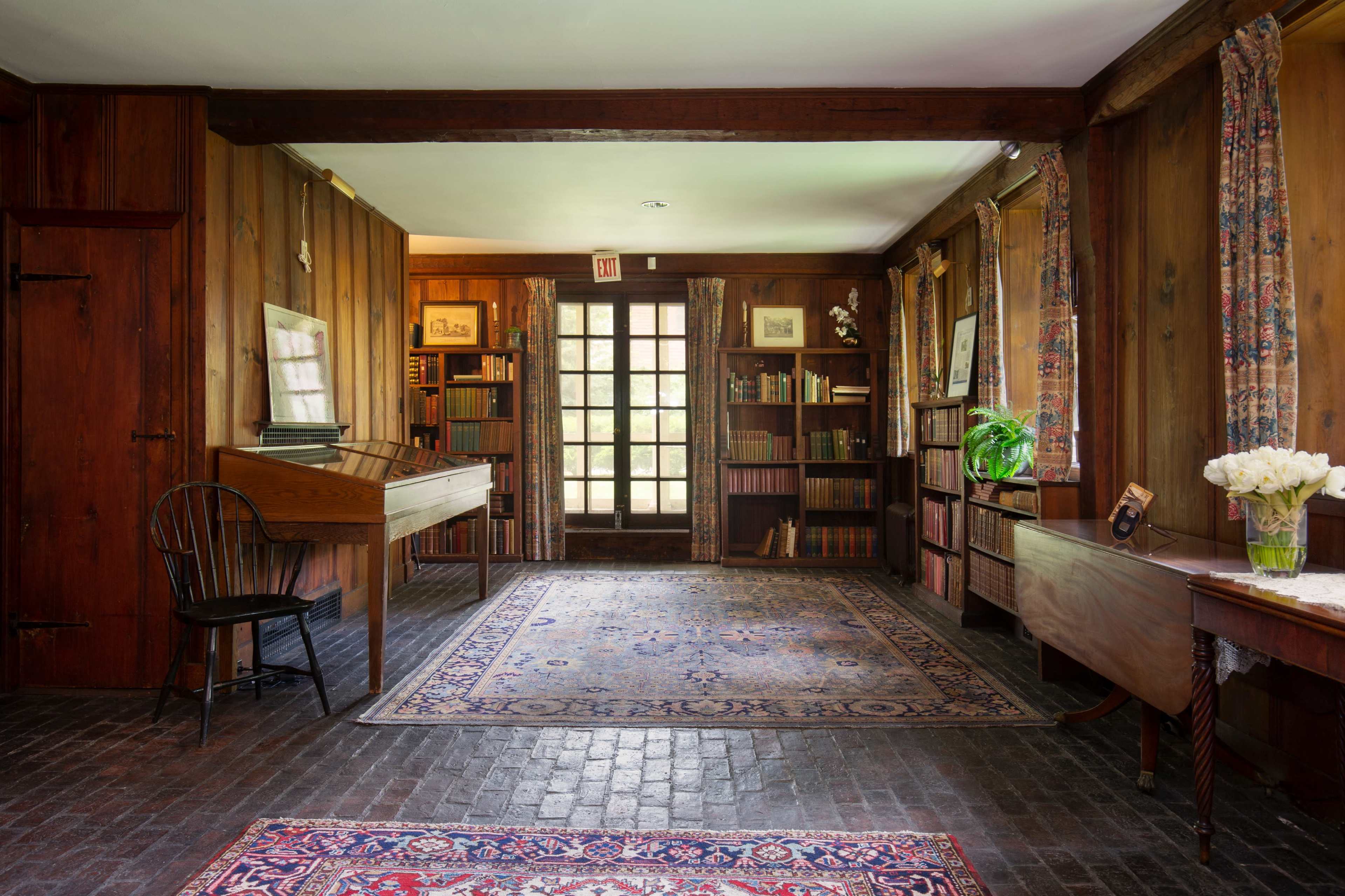 A wooden-paneled room with bookshelves lining the walls, a writing desk, a chair, and a large area rug in the center.