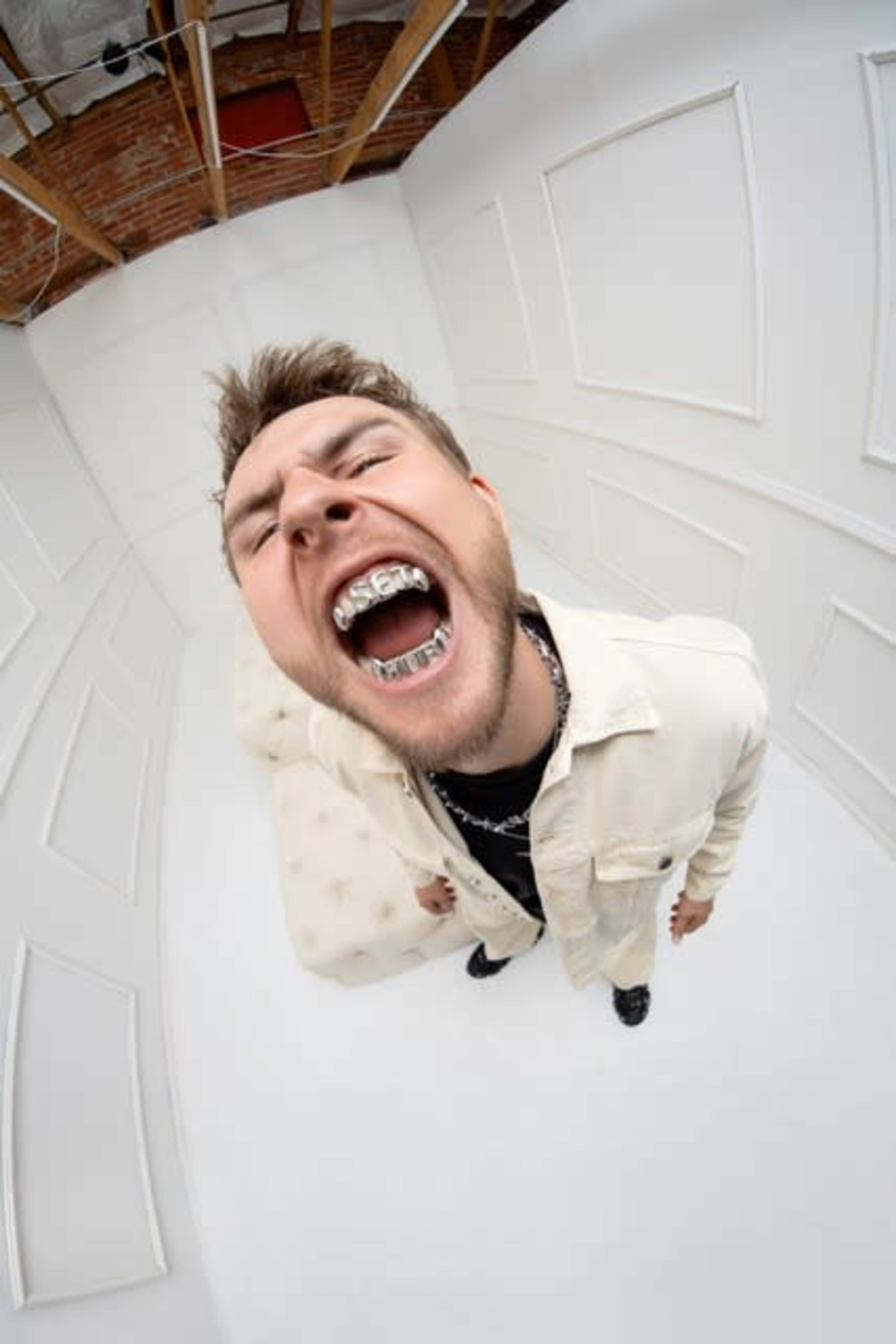 A person wearing a white outfit and dental grills is standing in a stark white room with a high ceiling, displaying an open-mouth expression.