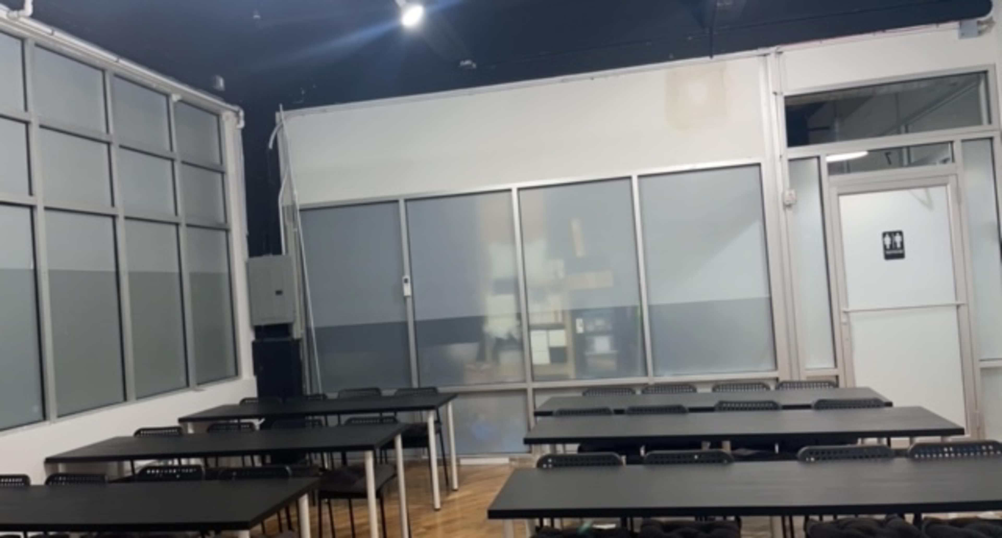 A classroom with rows of black tables and chairs arranged in a spacious room, featuring a glass wall and a restroom door.