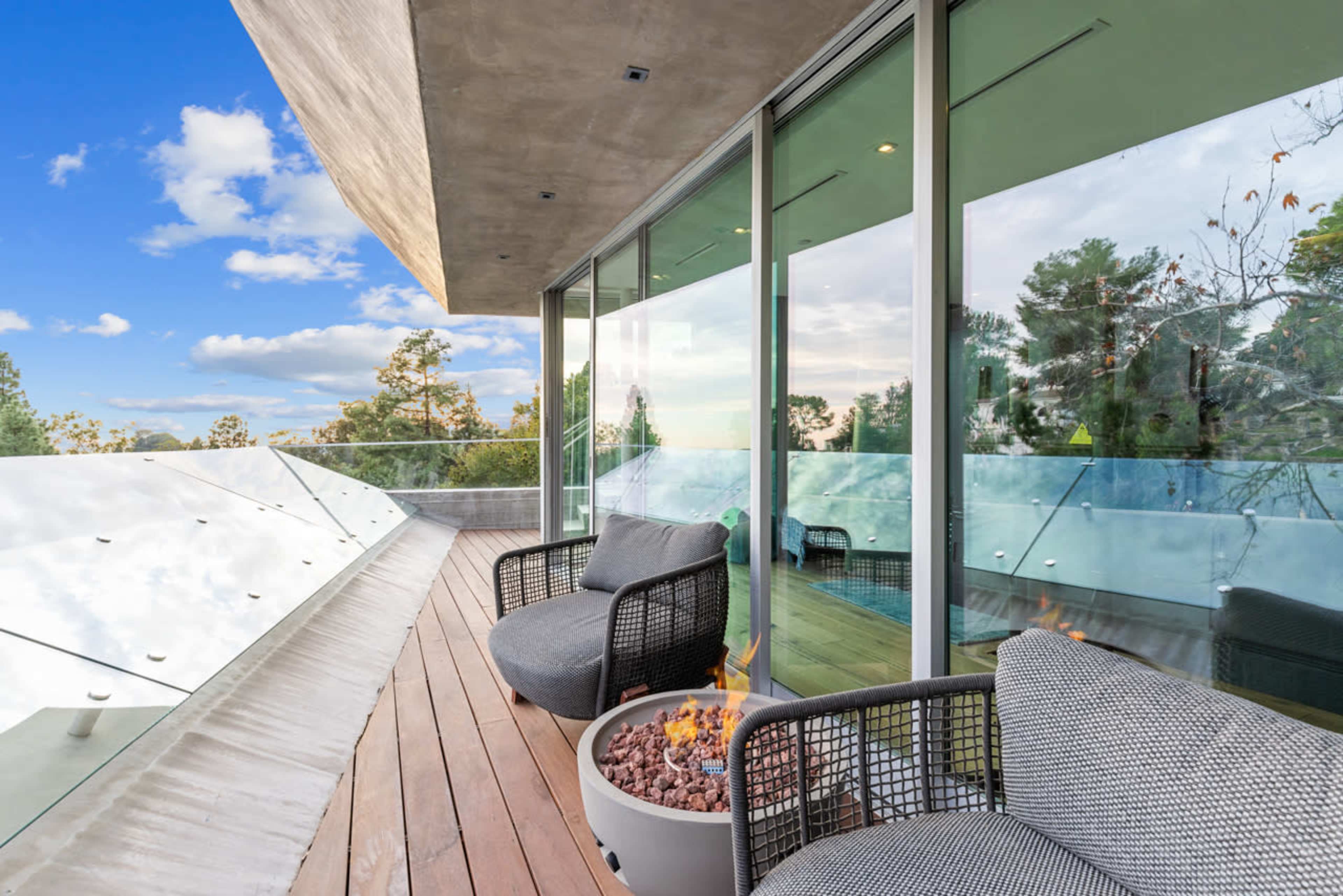 A modern balcony features two chairs and a fire pit, with glass railings and a view of trees under a blue sky.