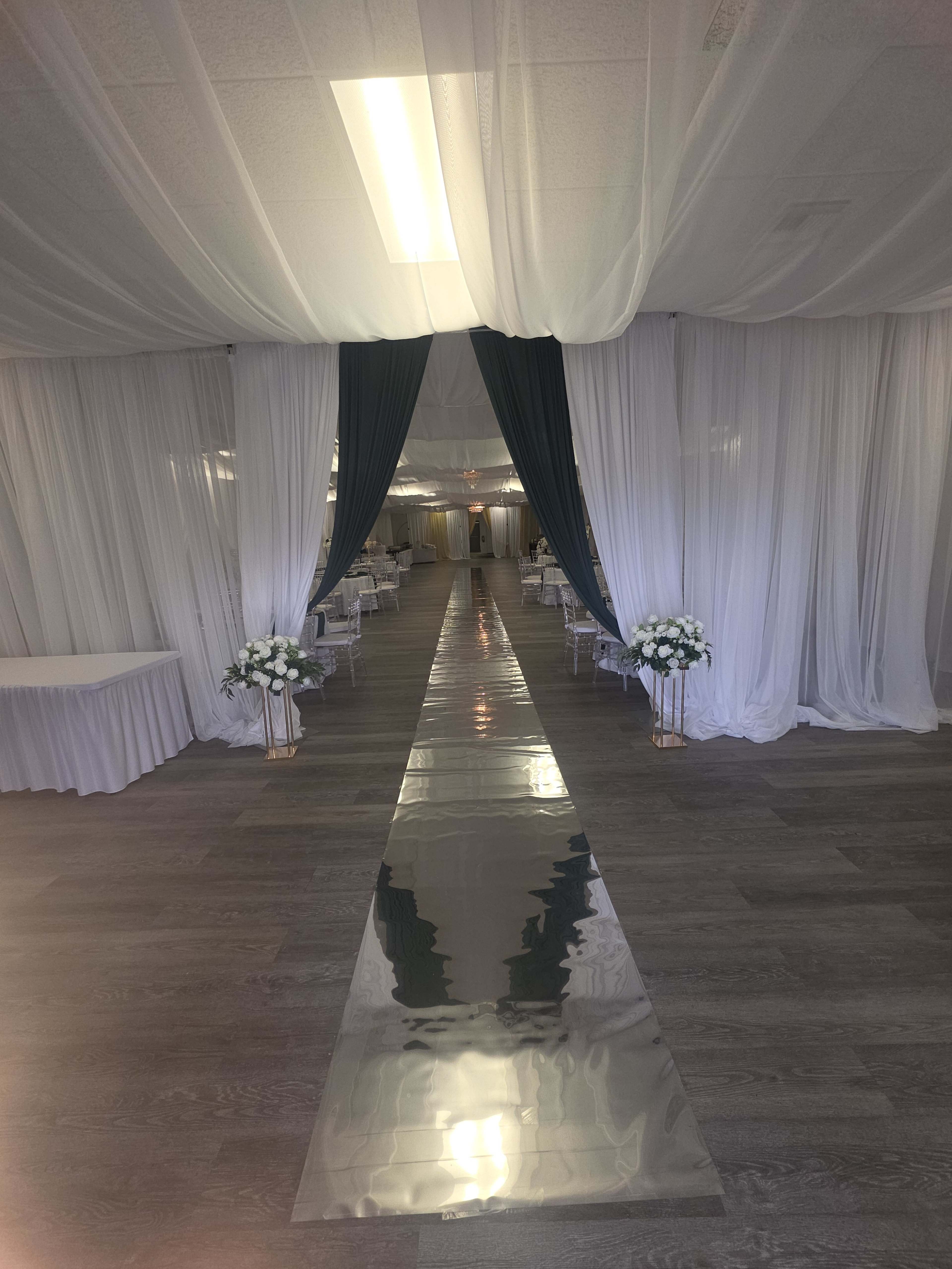 The image shows a spacious, decorated hallway with sheer white drapes and teal accents, leading to a set of tables arranged for an event.