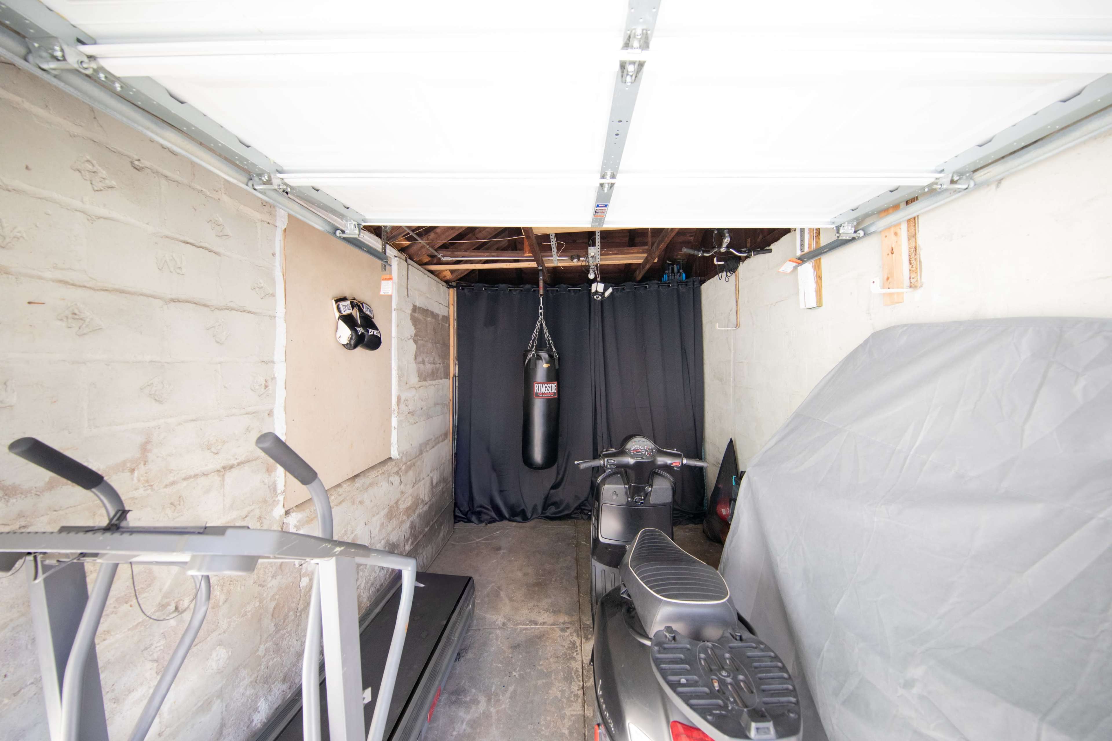 The image shows an empty garage with a treadmill, a motorcycle, and a heavy bag suspended from the ceiling, all set against a backdrop of unfinished walls.