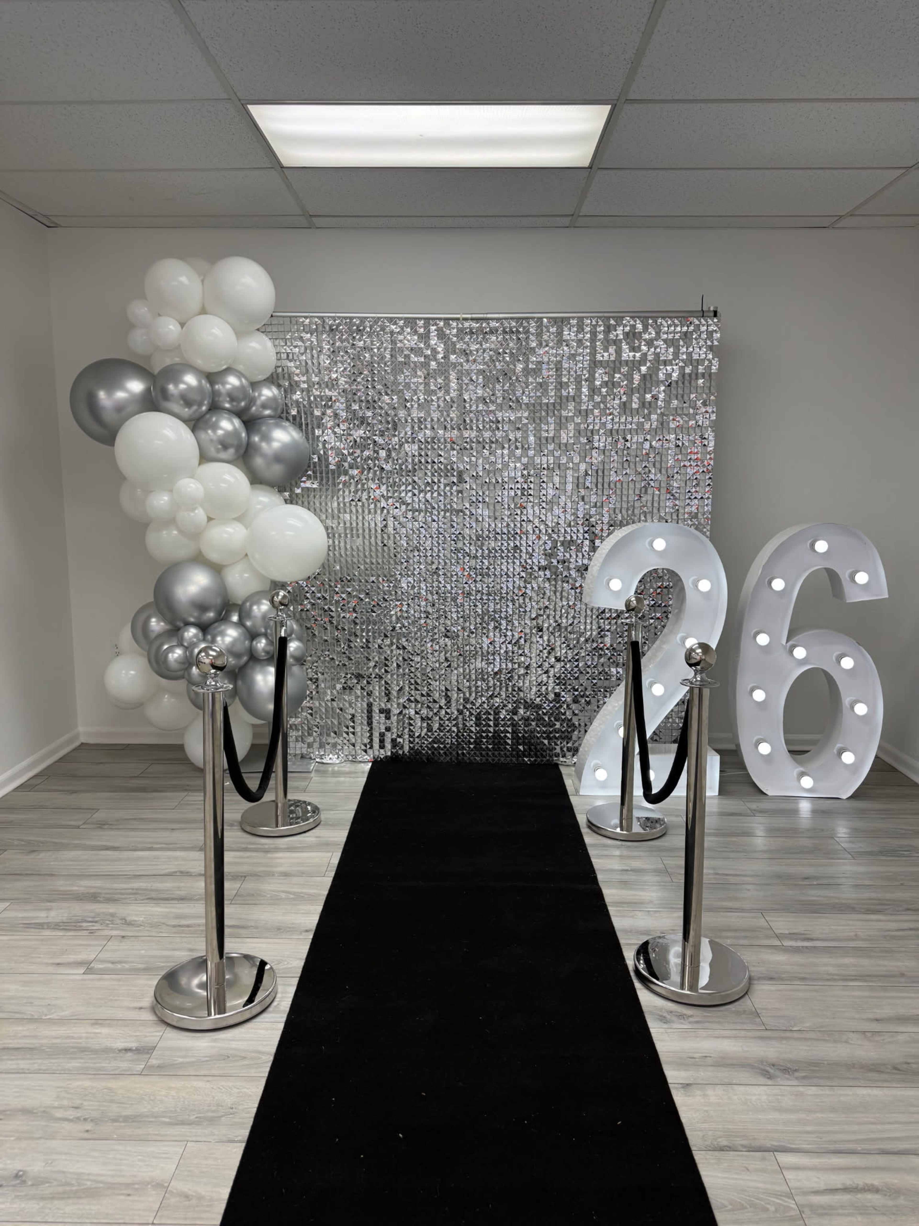 The image shows a black carpet leading to a sequined silver backdrop, with large white and silver balloons on one side and illuminated numbers "26" on the other side.