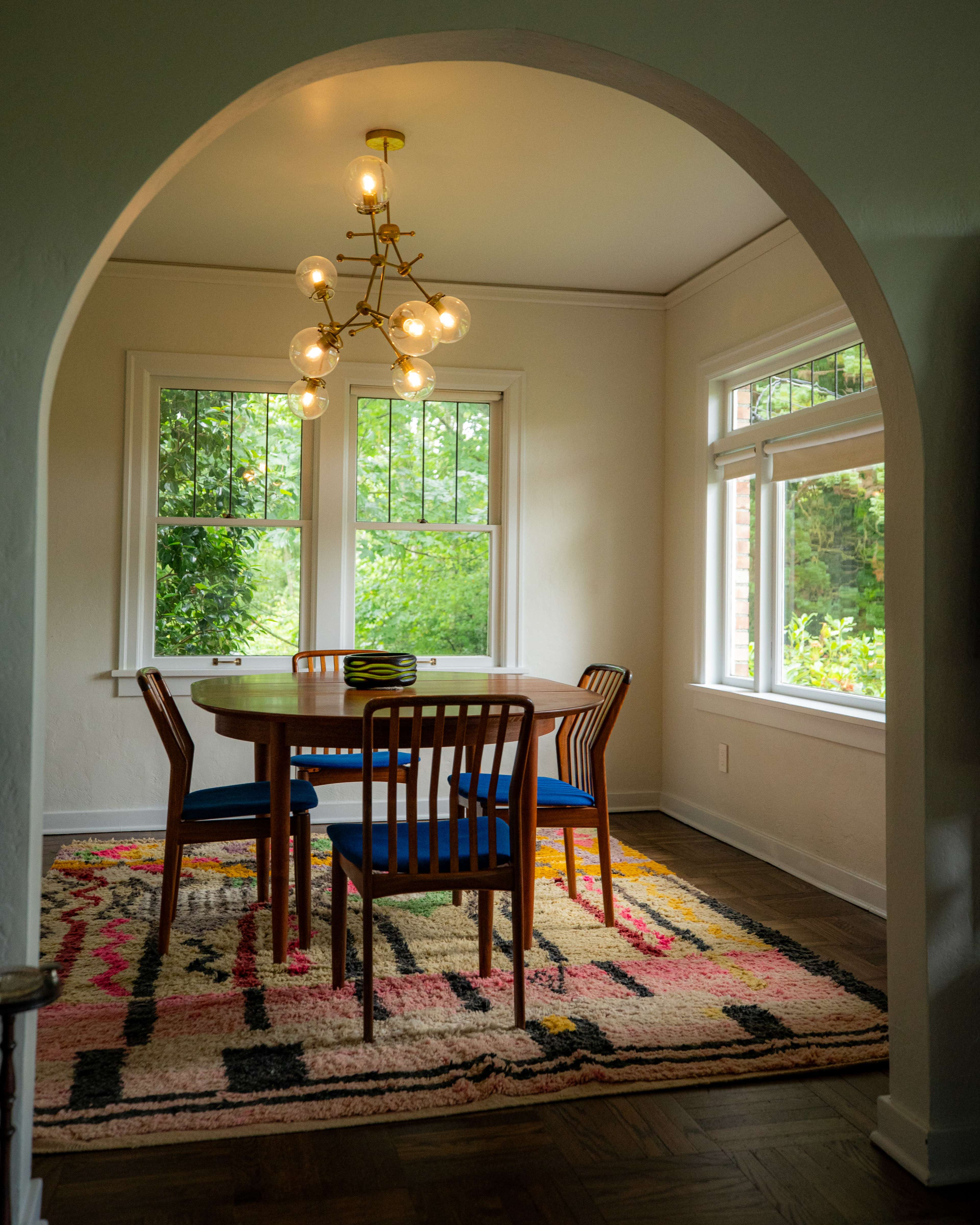 A round wooden dining table is surrounded by four wooden chairs on a colorful patterned rug in a well-lit room with large windows.