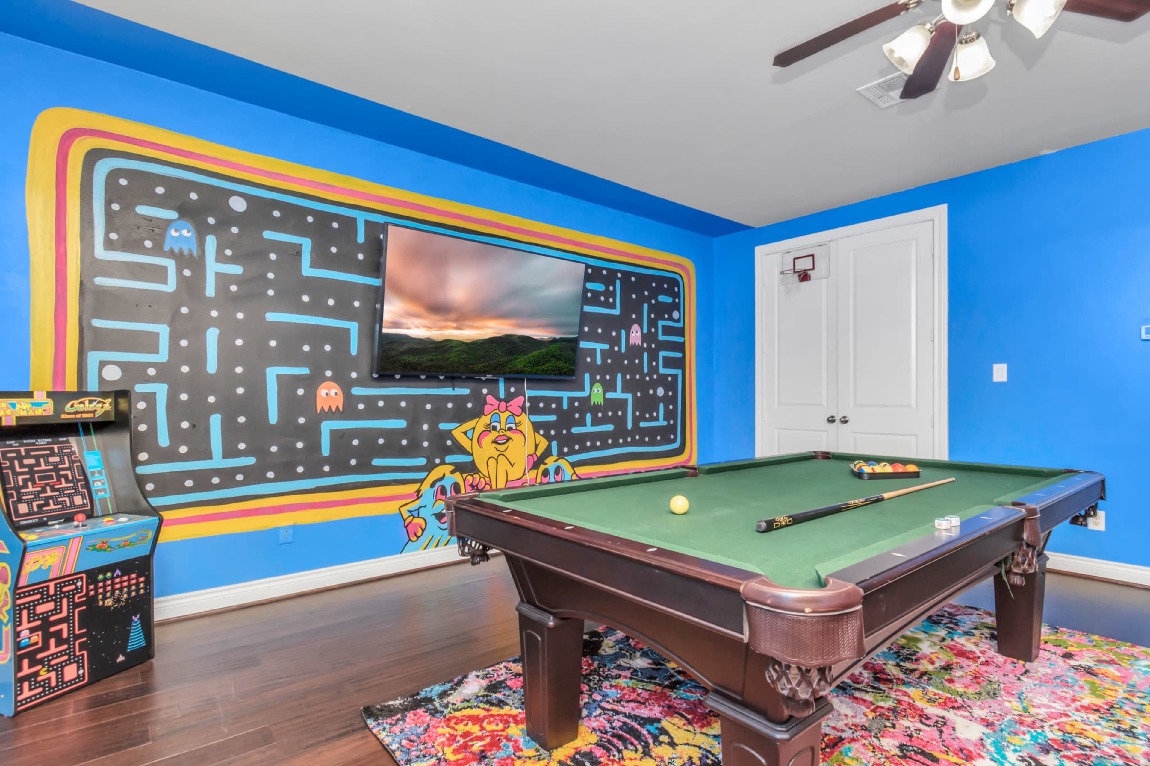 A pool table is set in a room featuring a vibrant blue wall with a retro arcade game and a large television mounted above a colorful Pac-Man mural.
