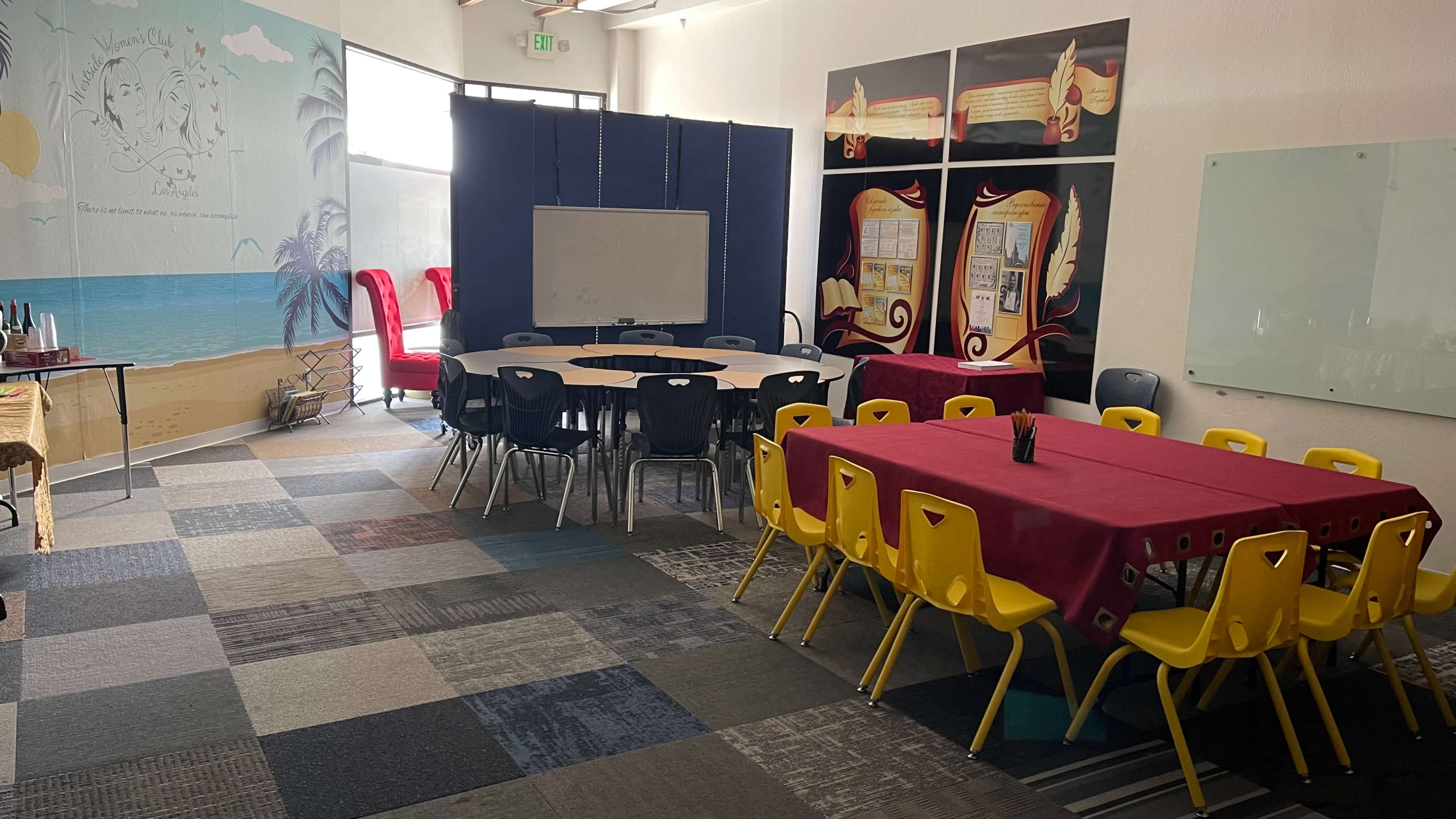 A brightly colored room with several round tables surrounded by yellow chairs, a large whiteboard, and a mural depicting a beach scene.