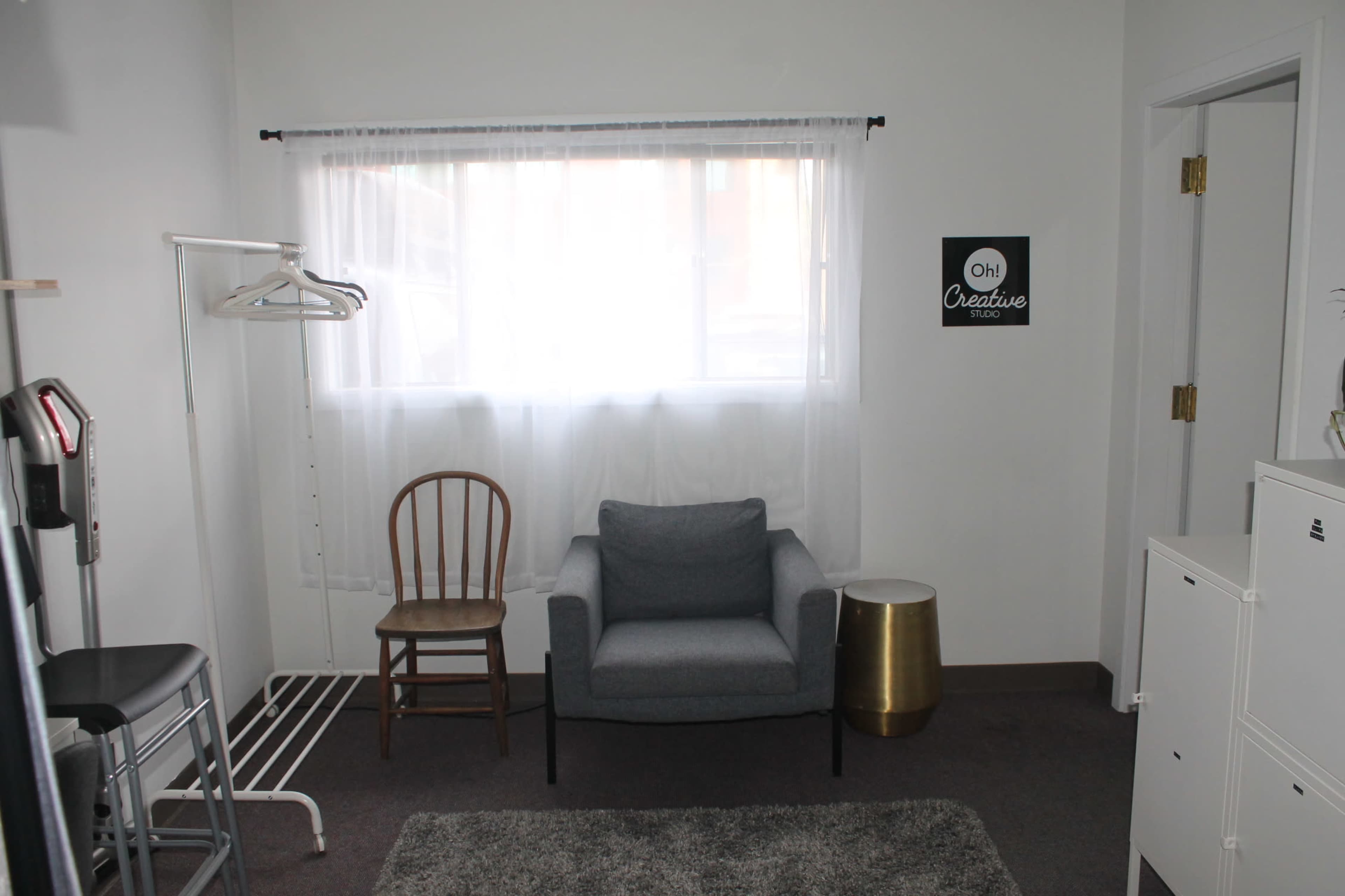 A small room with a chair, a gray couch, a clothing rack, and a window with sheer curtains.