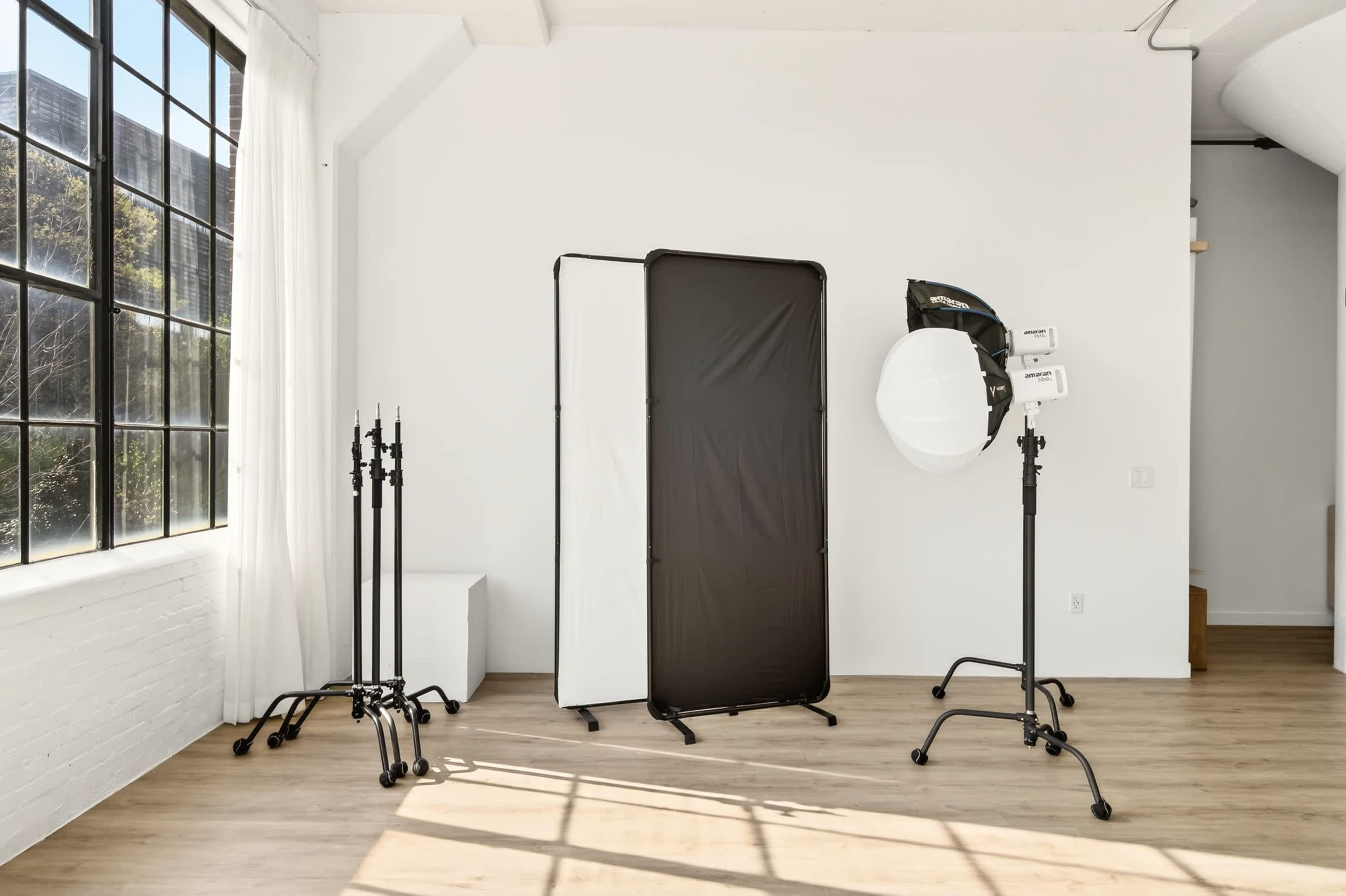 The image shows a spacious photography studio with a large window, a backdrop setup, and various lighting equipment on stands.