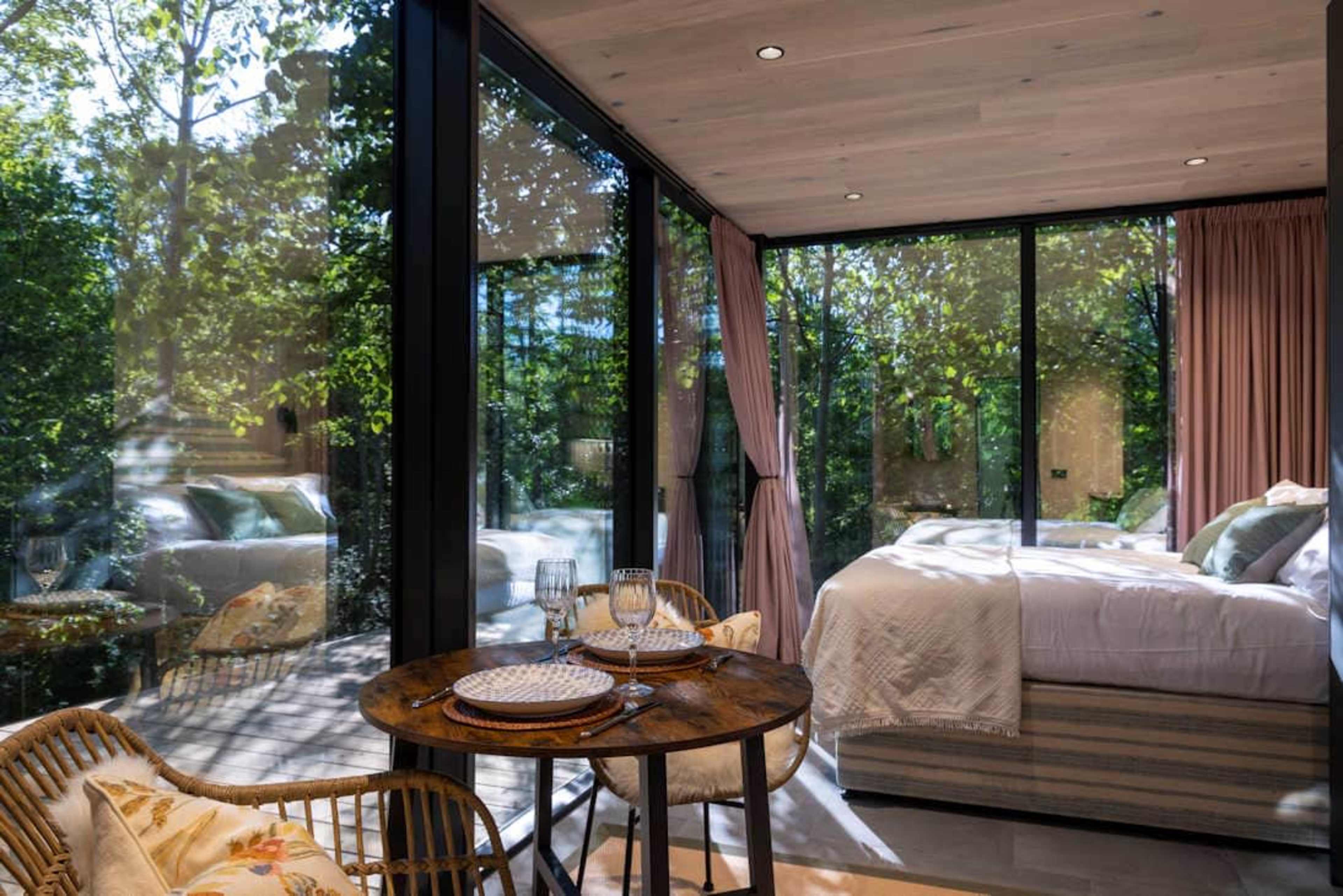 A modern bedroom features large windows overlooking a lush garden, with a dining table set for two in the foreground.