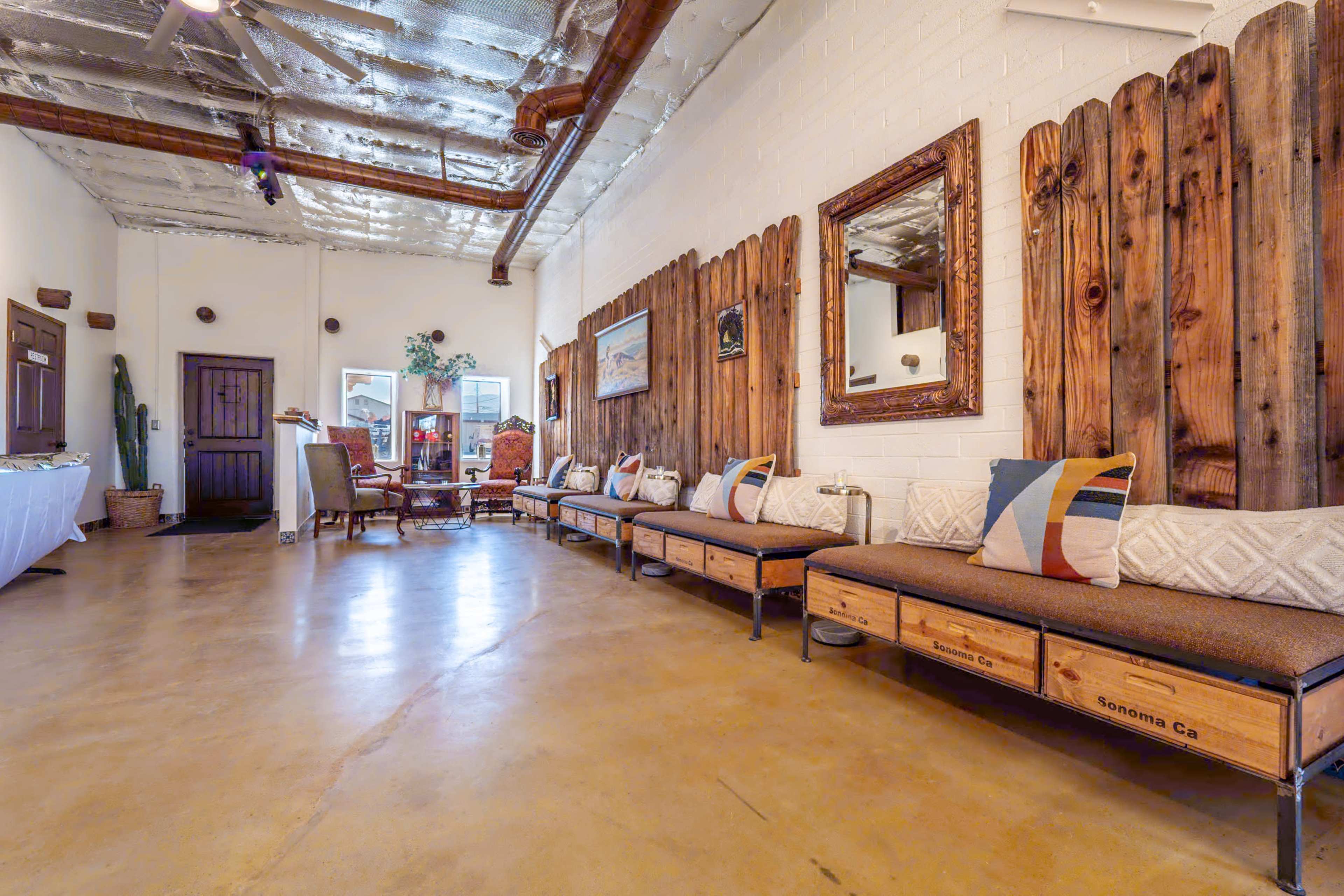 The image shows a spacious interior with wooden benches along a wall, adorned with decorative pillows, and featuring rustic wooden paneling and a mirror across from a seating area.