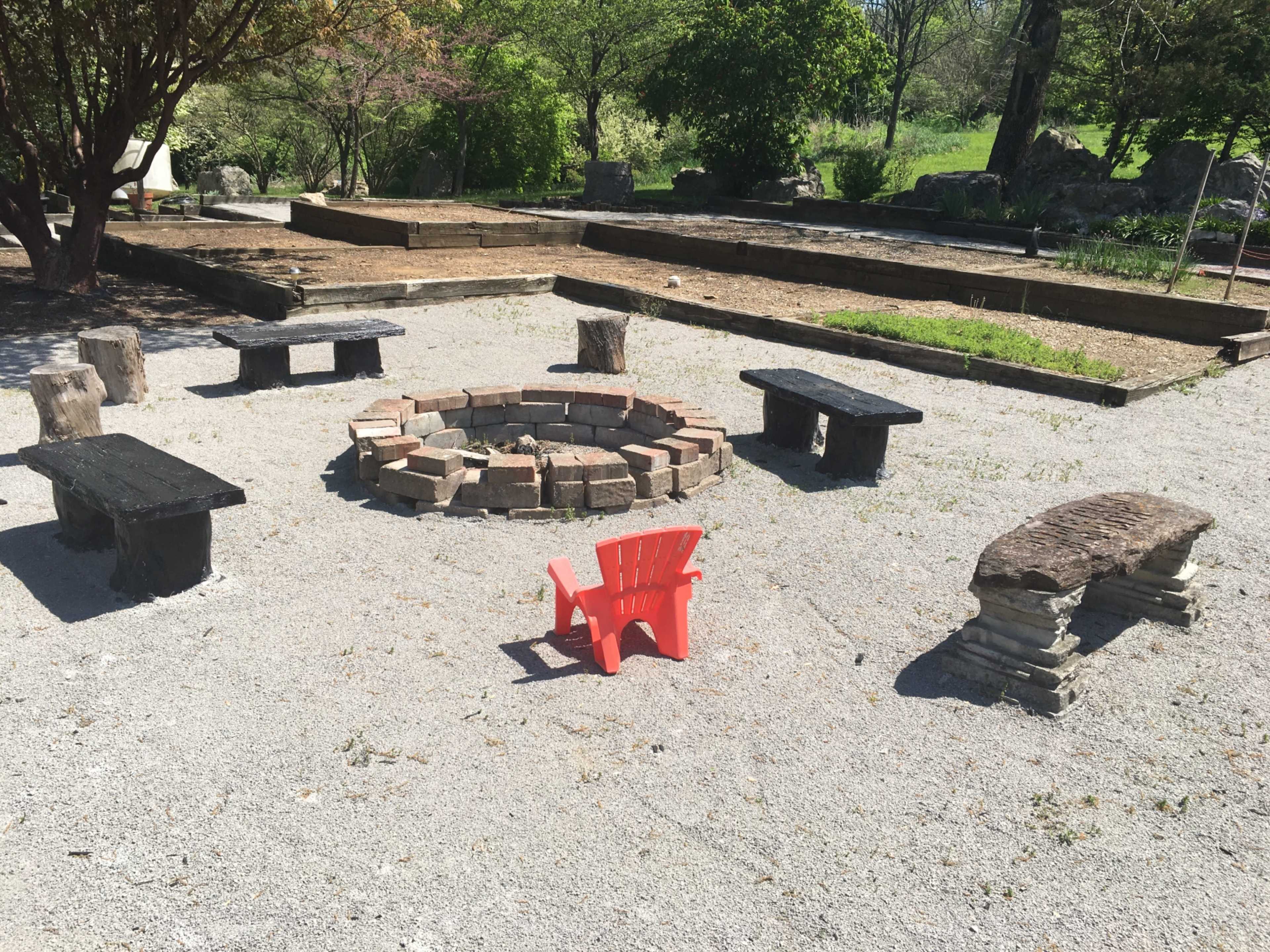 A circular stone fire pit is surrounded by several wooden benches and a single red Adirondack chair in a gravel area.