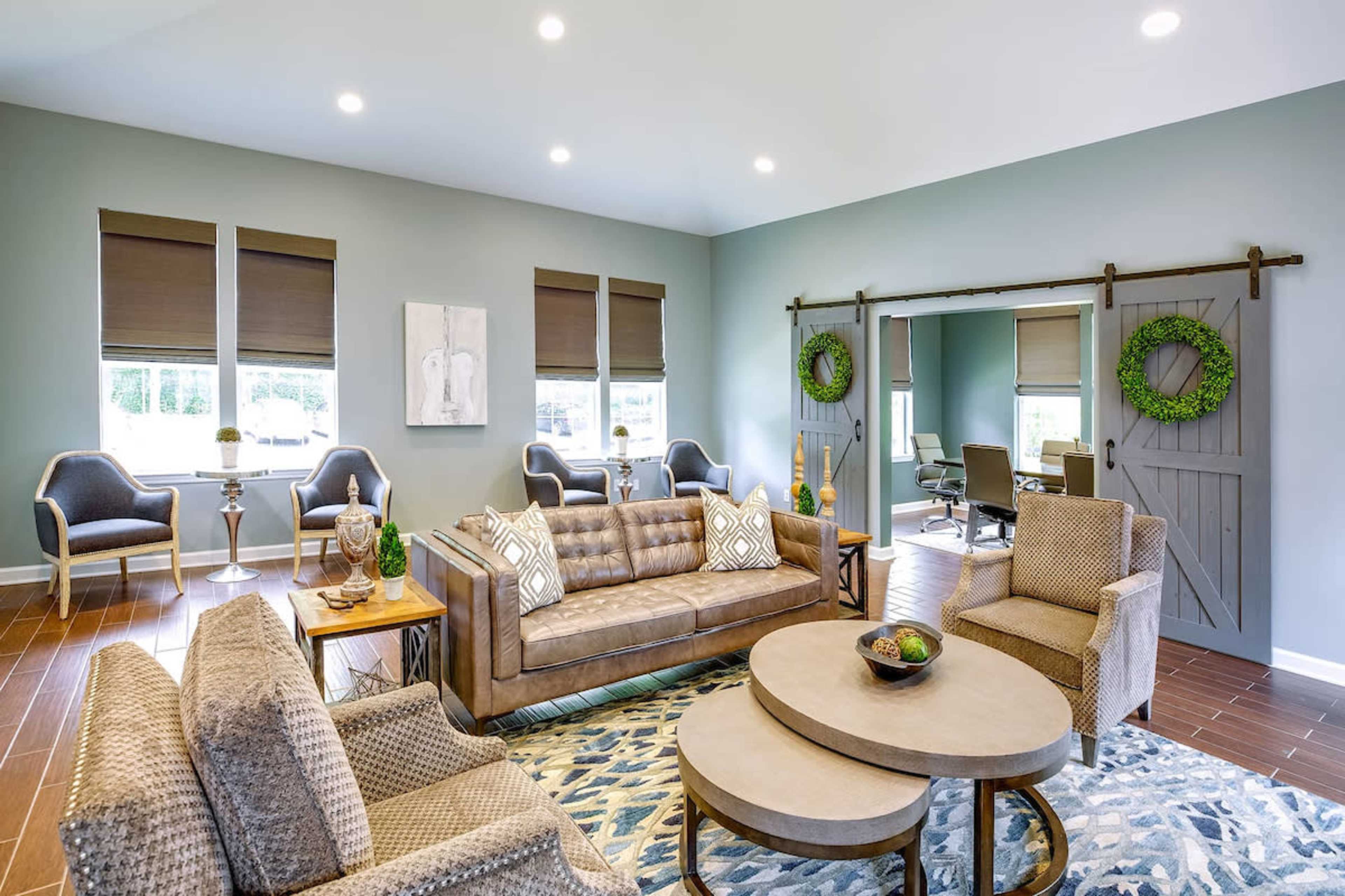 The image shows a modern living room with a brown sofa, patterned chairs, a large area rug, and windows adorned with brown shades, along with a sliding barn door.
