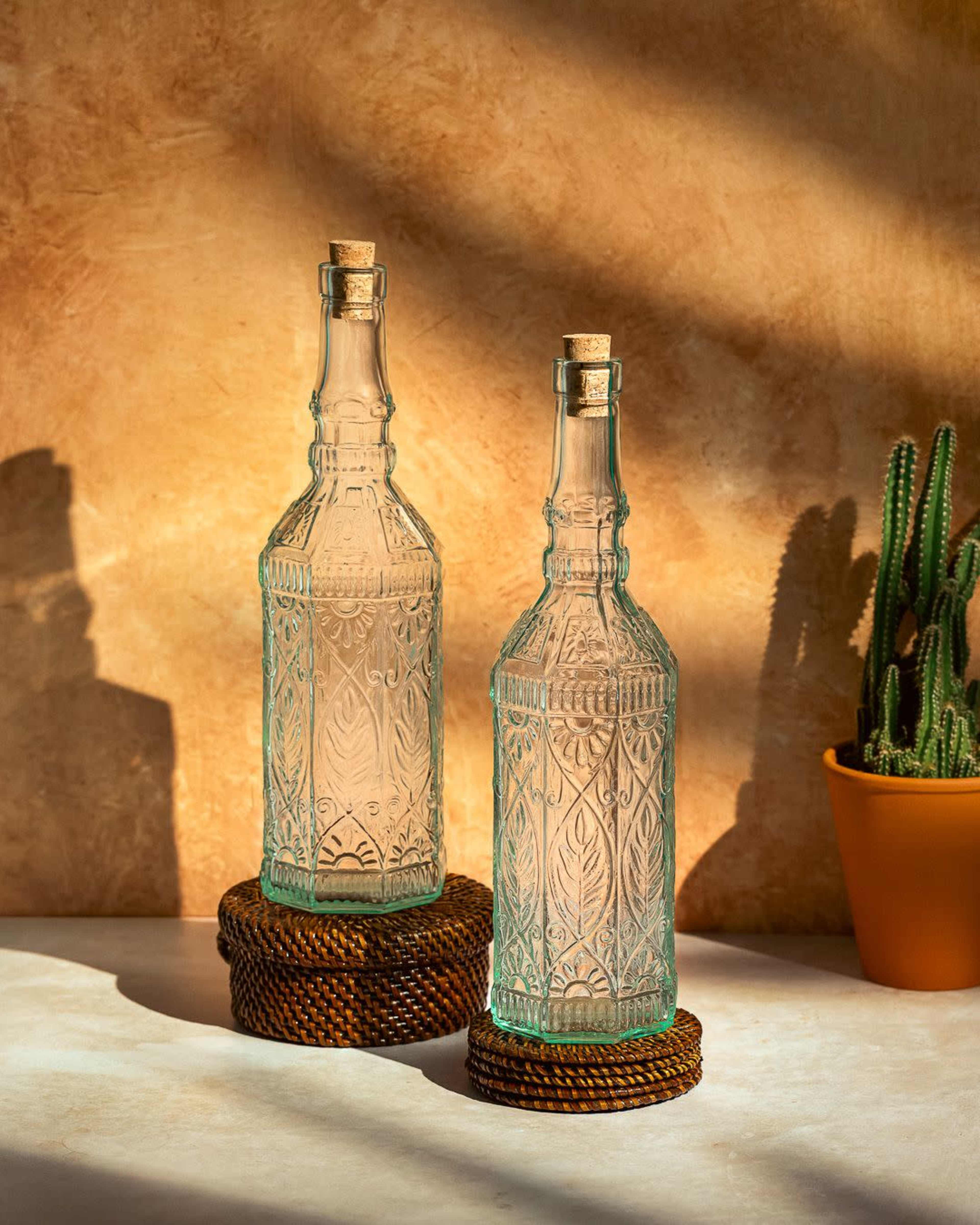 Two decorative glass bottles with cork stoppers are positioned on a woven coaster, set against a textured, warm-toned background with a small potted cactus nearby.