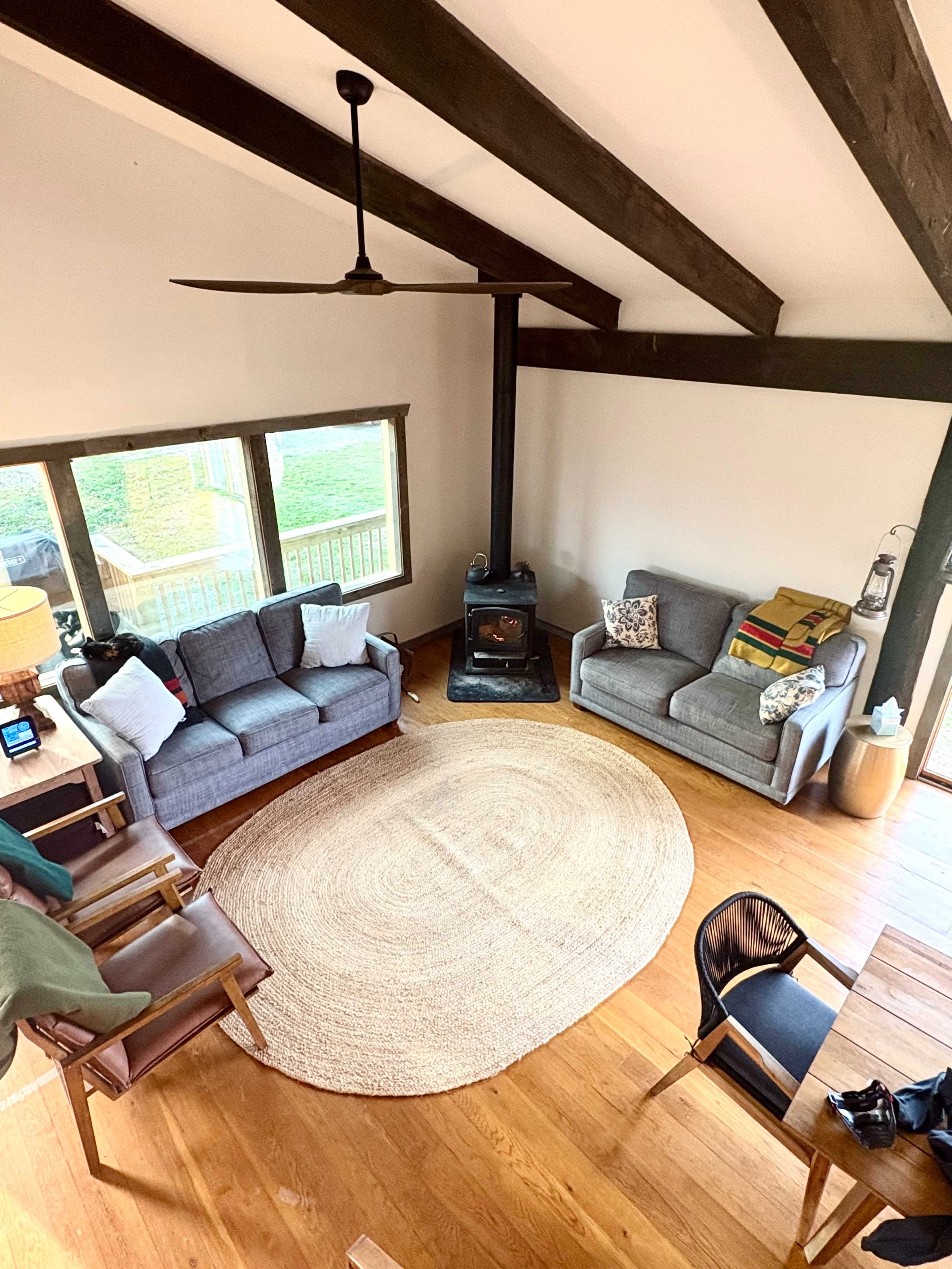 The image shows a cozy living room with two gray sofas, wooden beams on the ceiling, a circular rug on the wooden floor, and a black wood stove near large windows.
