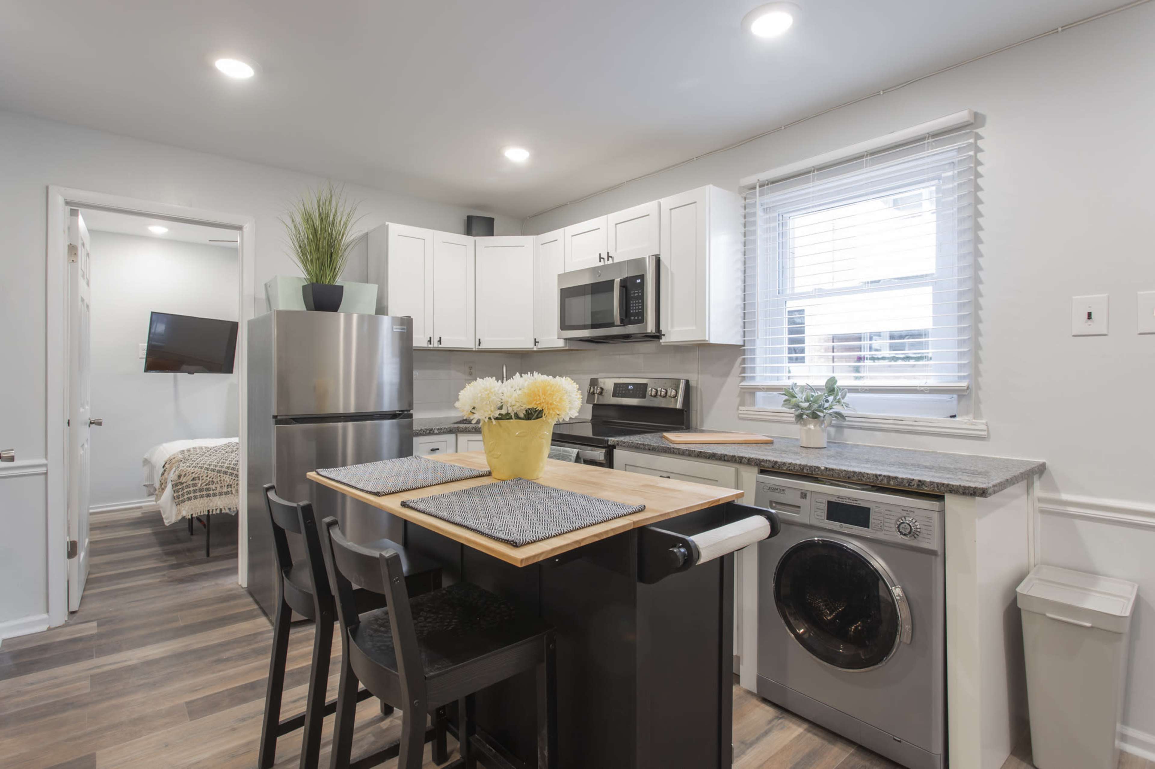 A modern kitchen features a stainless steel refrigerator, a washer, and a wooden dining table with four chairs.