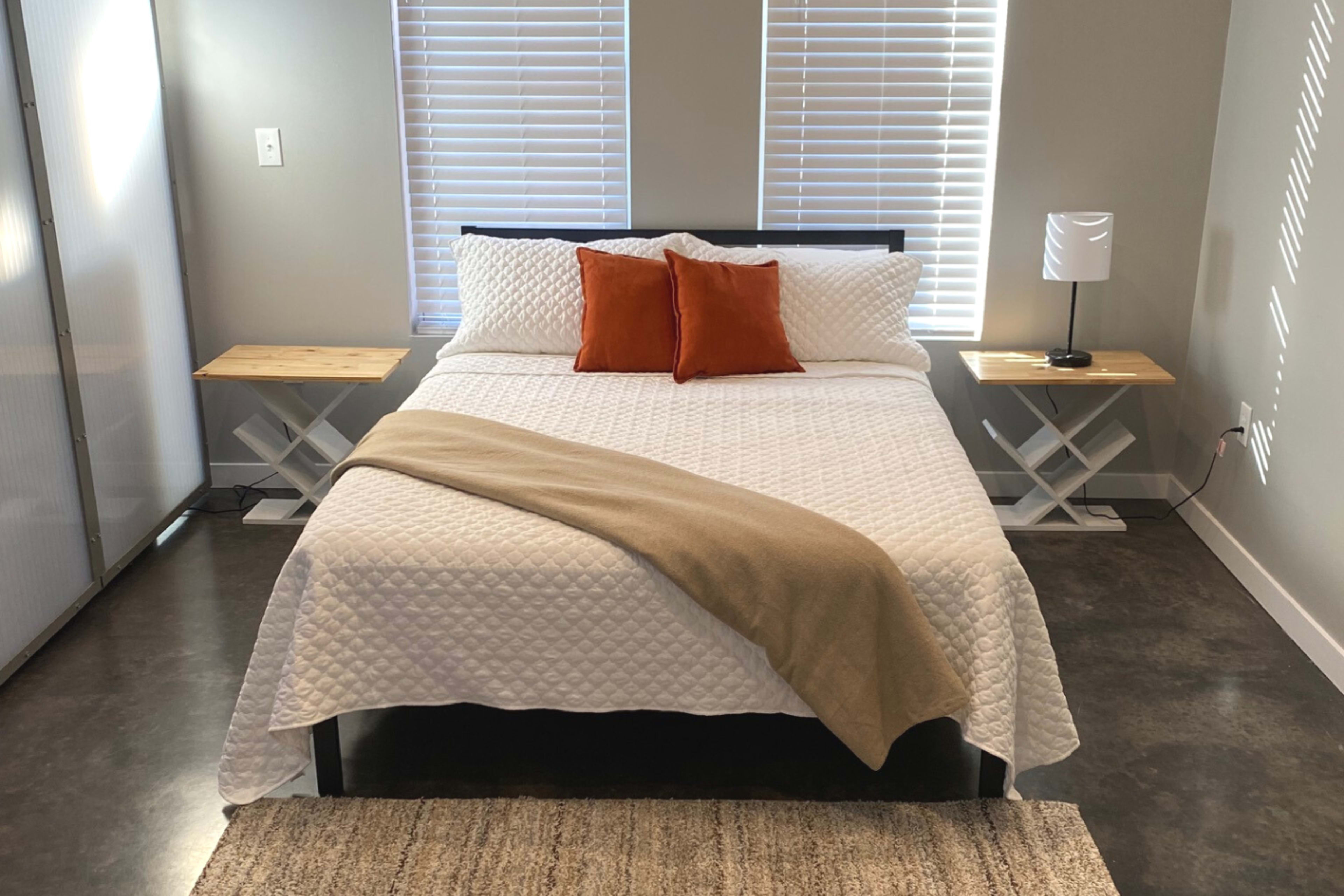 The image shows a neatly arranged bedroom with a bed featuring white bedding and two decorative pillows, flanked by wooden side tables and a lamp.