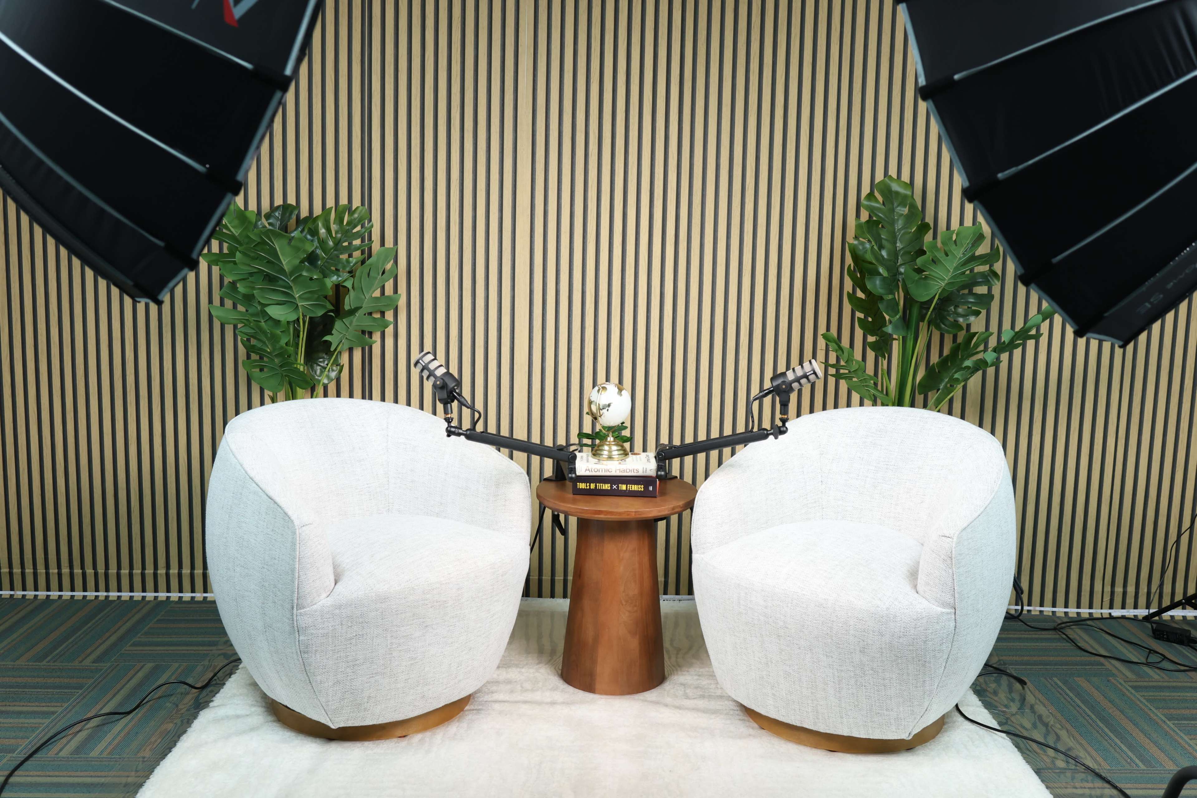 The image shows two white round chairs positioned opposite each other on a light rug, with a wooden table between them and two microphones attached to stands.