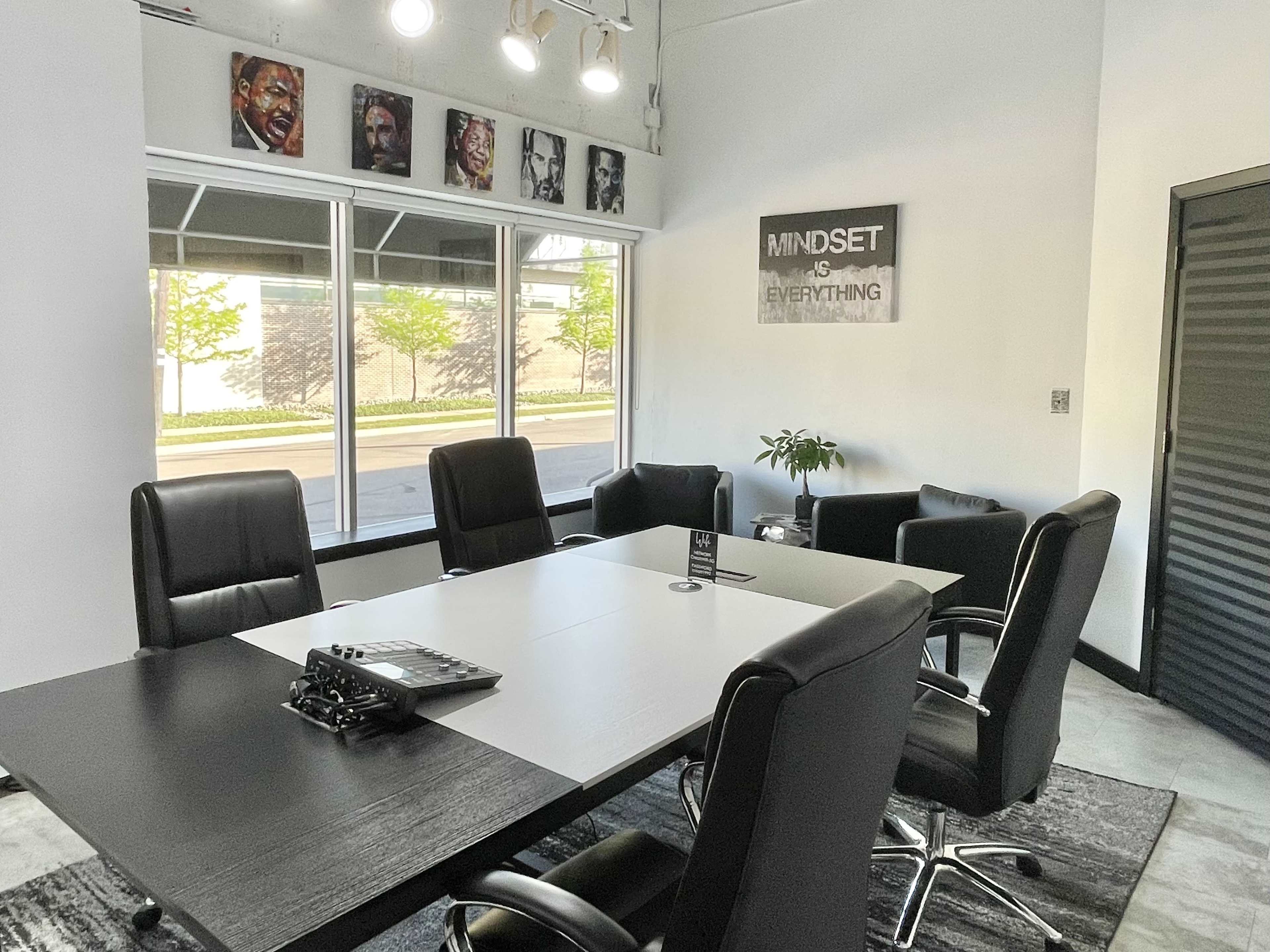 A modern conference room with a large table surrounded by black chairs, featuring windows that provide natural light and a poster on the wall that reads "MINDSET IS EVERYTHING."
