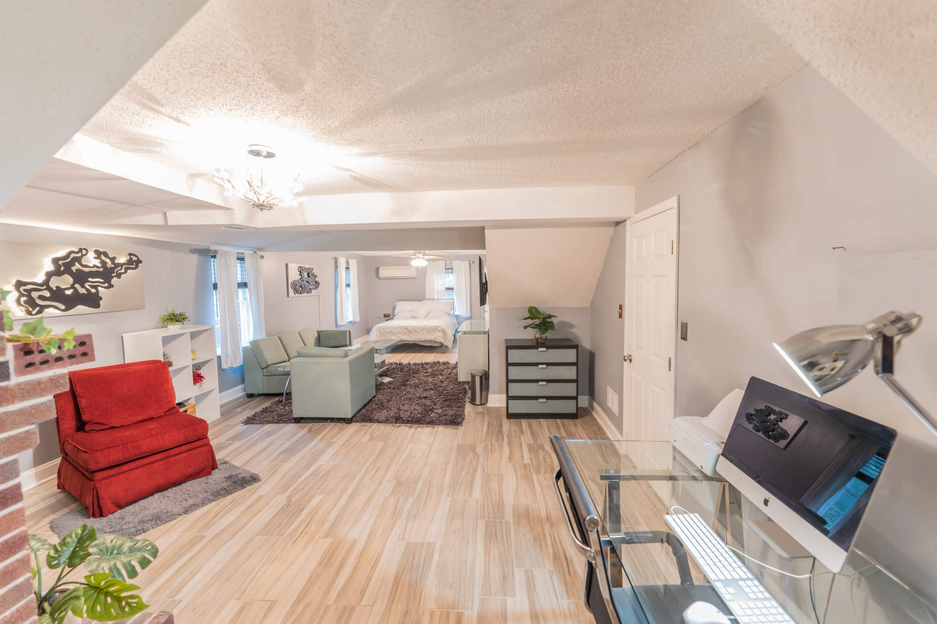 The image shows a well-lit open living space with a red chair, green armchairs, a glass desk, and a bed in the background.