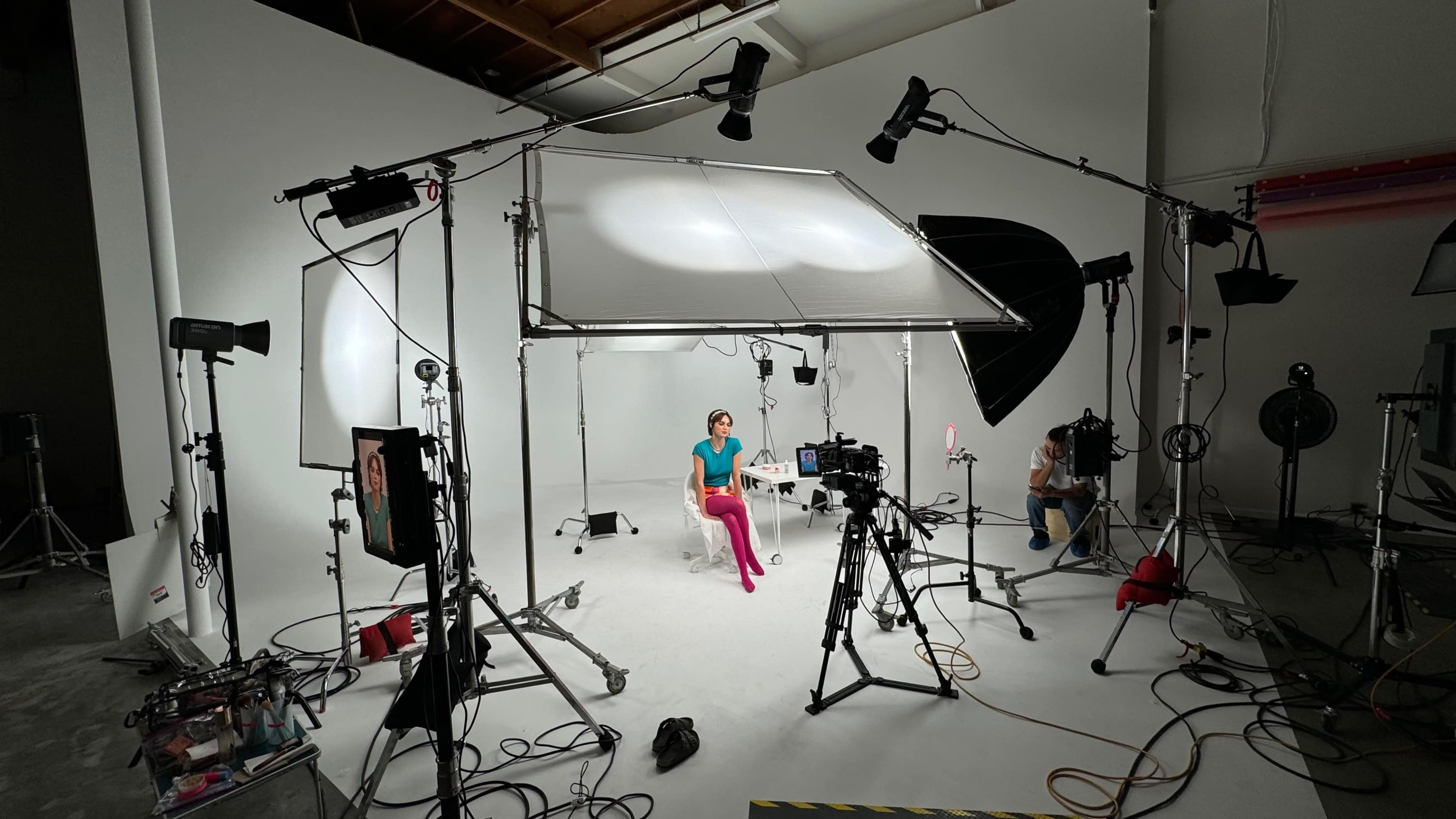 A studio setup features a seated model with bright pink pants, surrounded by various lighting equipment and cameras against a white backdrop.