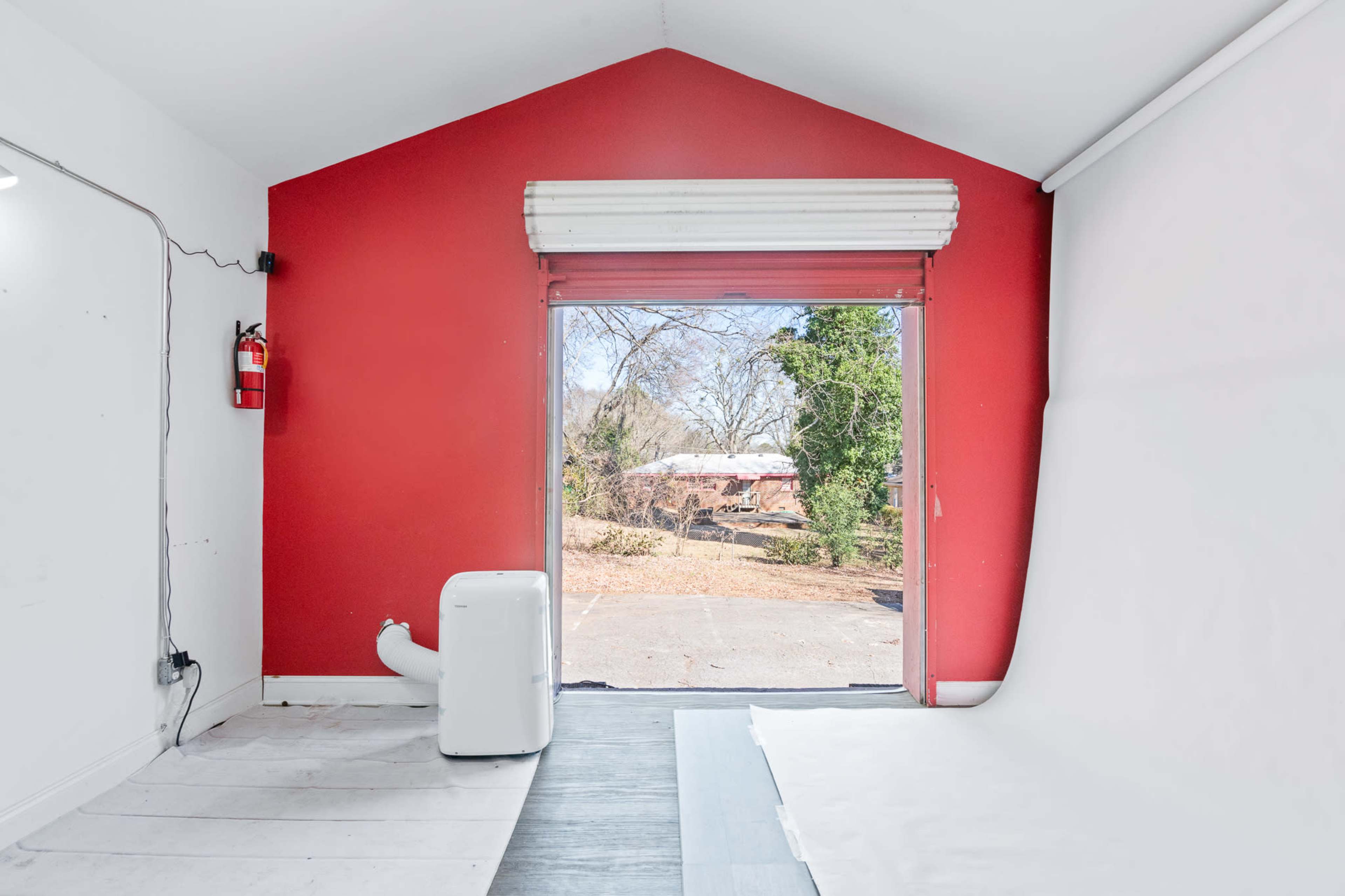 The image shows a room with a red wall, a white air conditioning unit on the floor, and a large opening that leads to an outdoor area.
