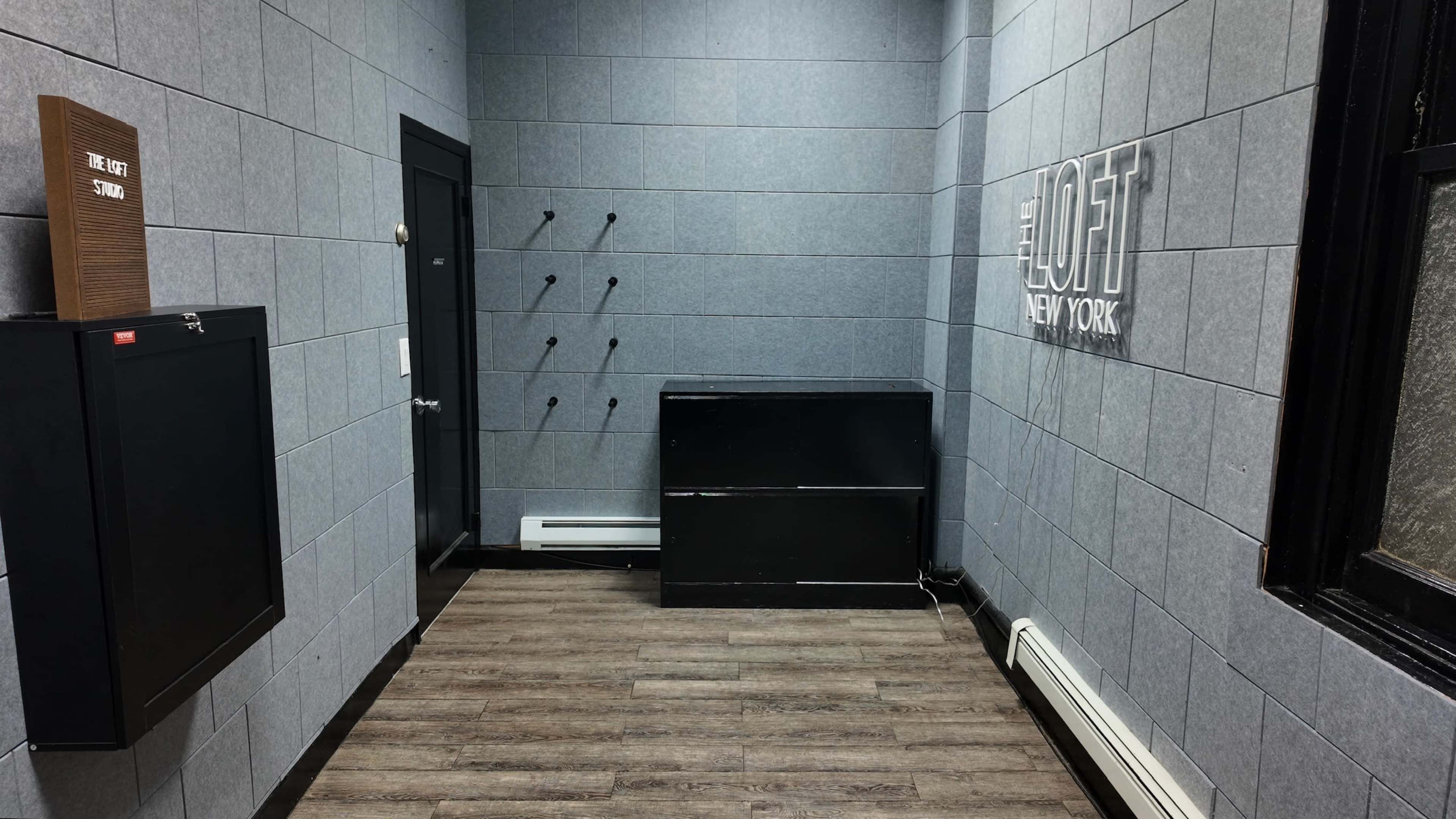 The image shows a small, minimalist hallway with gray walls, hardwood flooring, and a black storage unit.