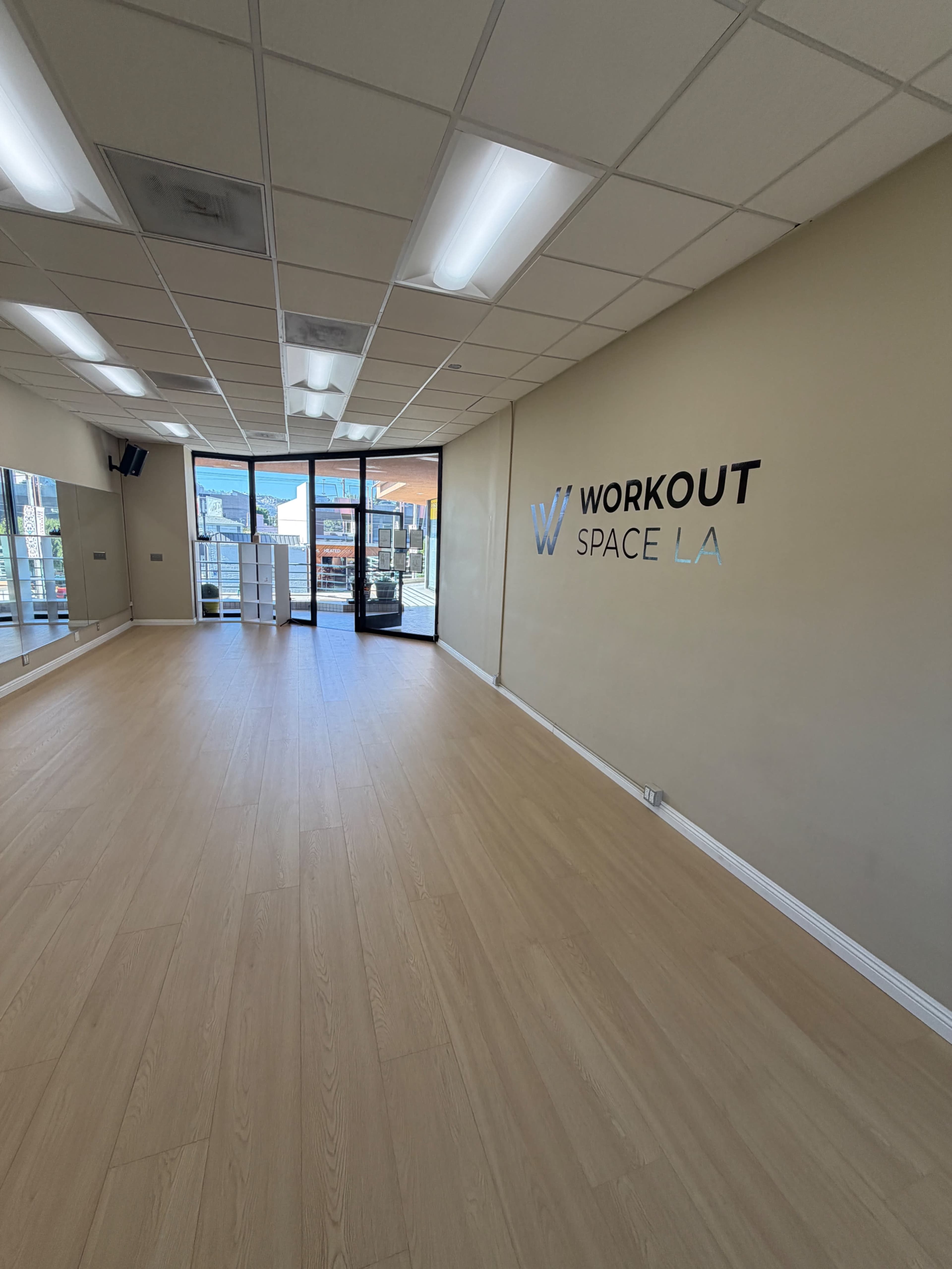 The image shows a spacious fitness studio with wooden flooring, large windows, and a sign reading "WORKOUT SPACE LA" on the wall.