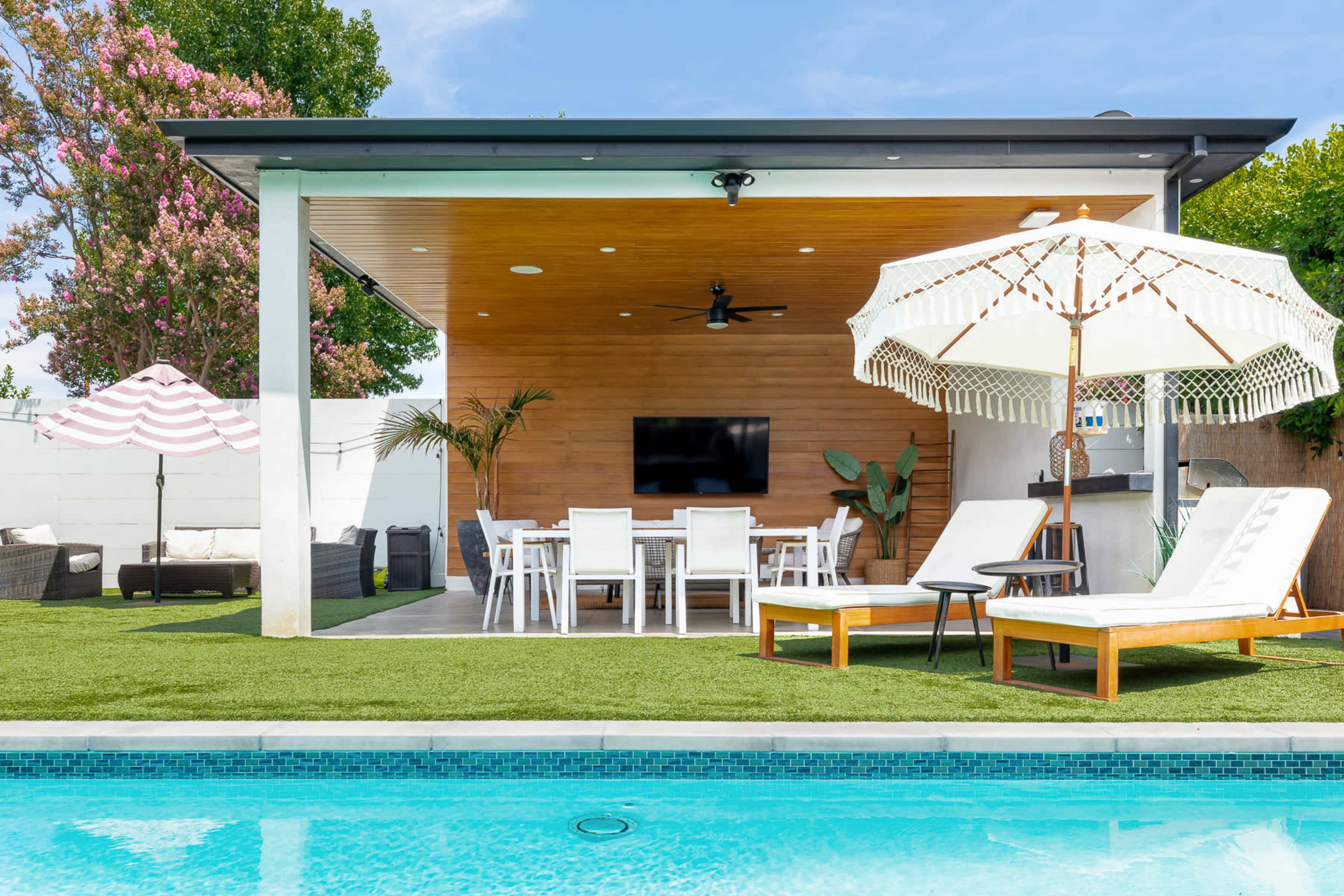A modern outdoor living area features a pool, lounge chairs, dining table, and a shaded patio with a television.