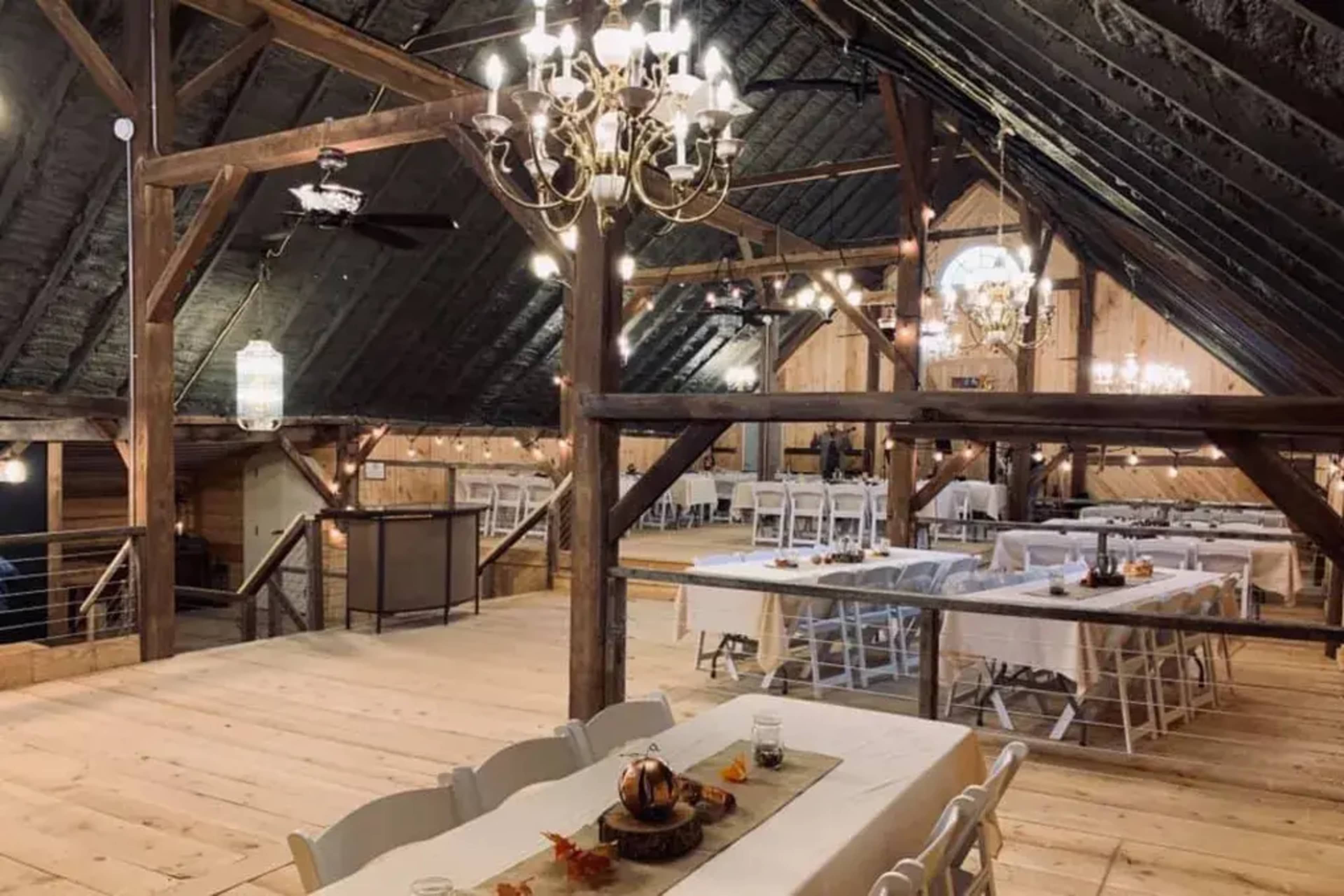 A spacious, rustic event space with wooden beams, white tables set for dining, and chandeliers hanging from the ceiling.
