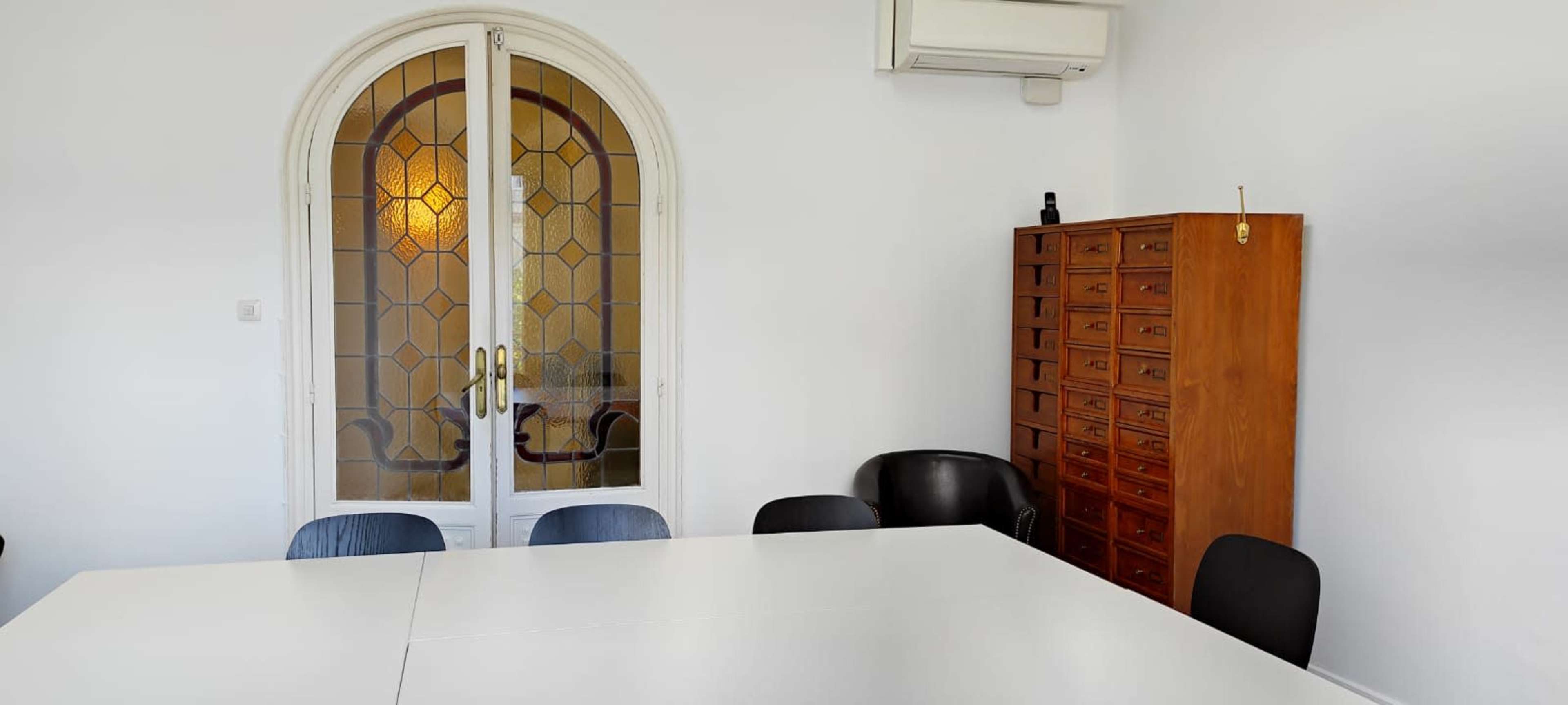 The image shows a conference room with a white table, several chairs, and a wooden cabinet against the wall, along with a set of decorative glass doors.